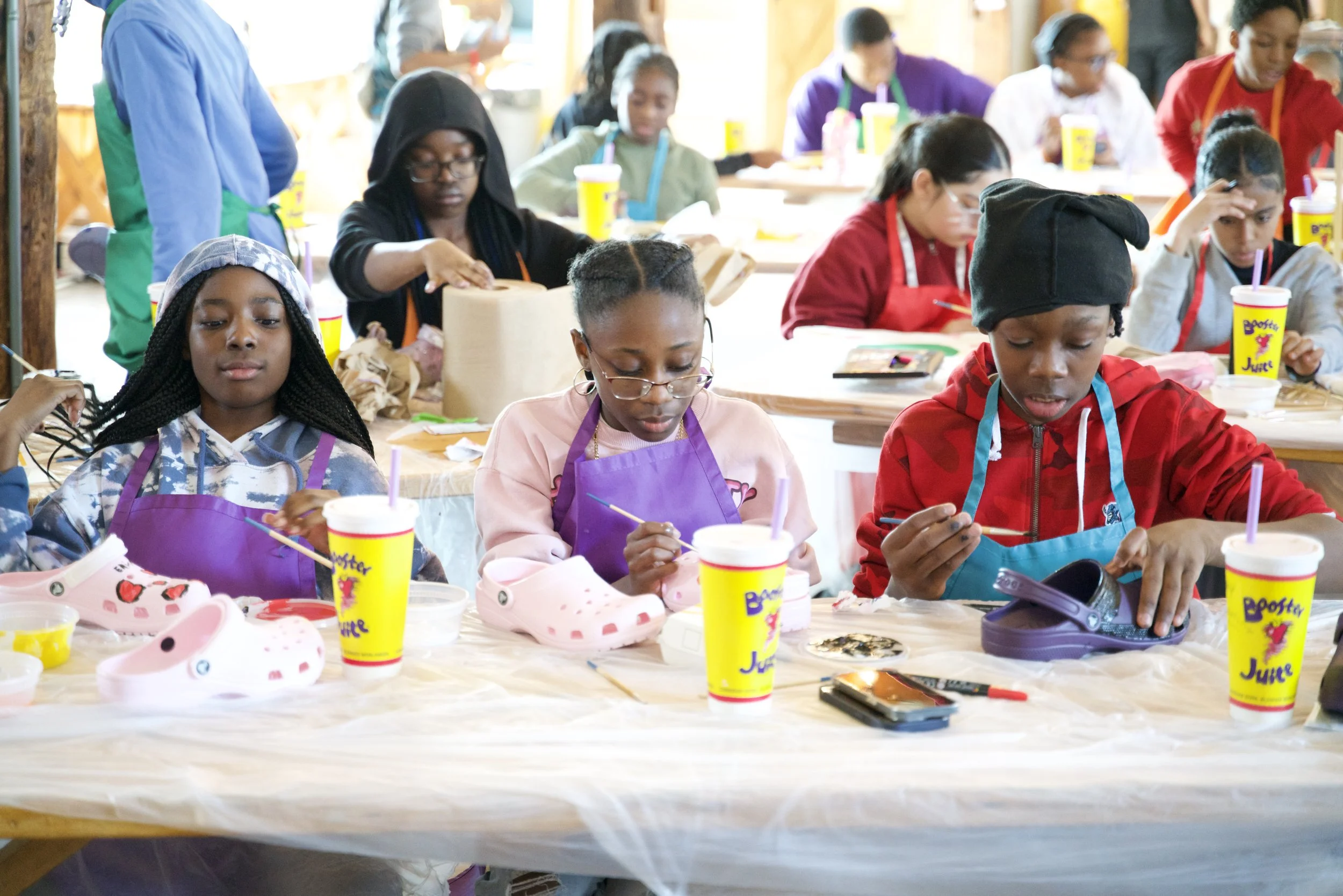 A group of children participating in a craft activity at a table in a bright and lively room. They are wearing aprons and hats, with supplies such as cups, shoes, and craft tools in front of them.