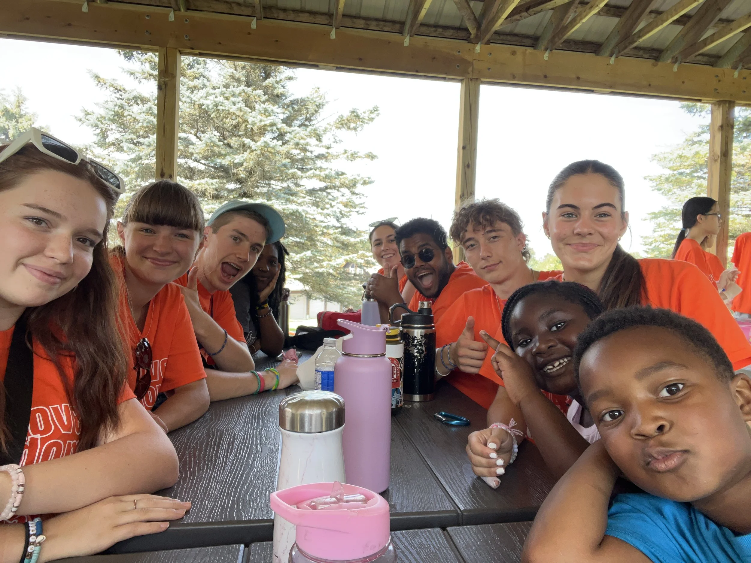 Group of smiling young people sitting together at a table outdoors under a wooden pavilion on a sunny day.