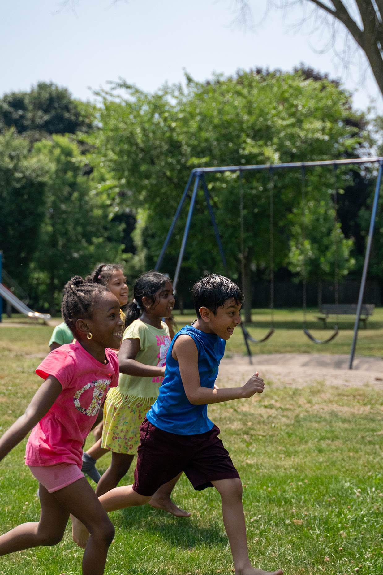 Group of children running together in a park on a sunny day, with a swing set and trees in the background.