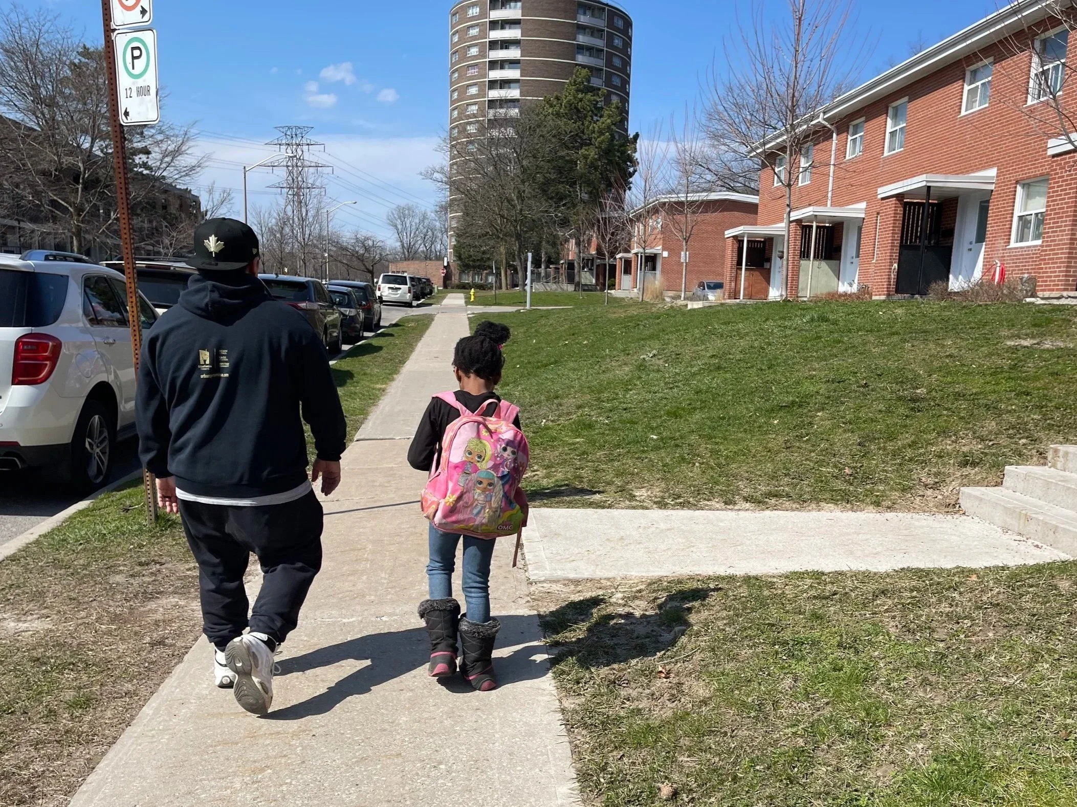 A person and a child walking on a sidewalk in a residential neighborhood with parked cars and brick row houses, a green grassy area, trees, and a tall apartment building in the background under a partly cloudy sky.