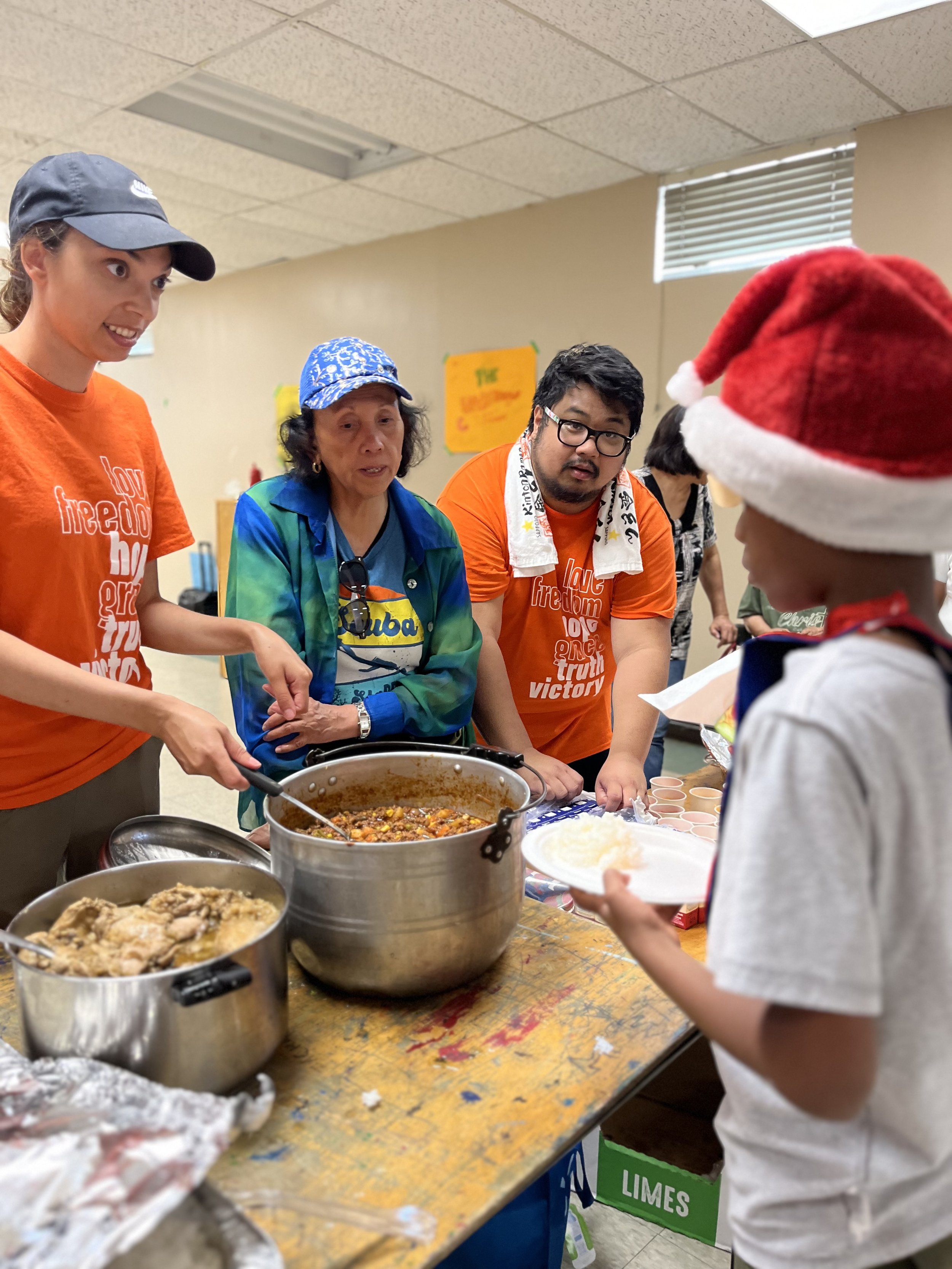People serving food at a community meal with pots of stew and plates of rice, with a person wearing a Santa hat in the foreground.