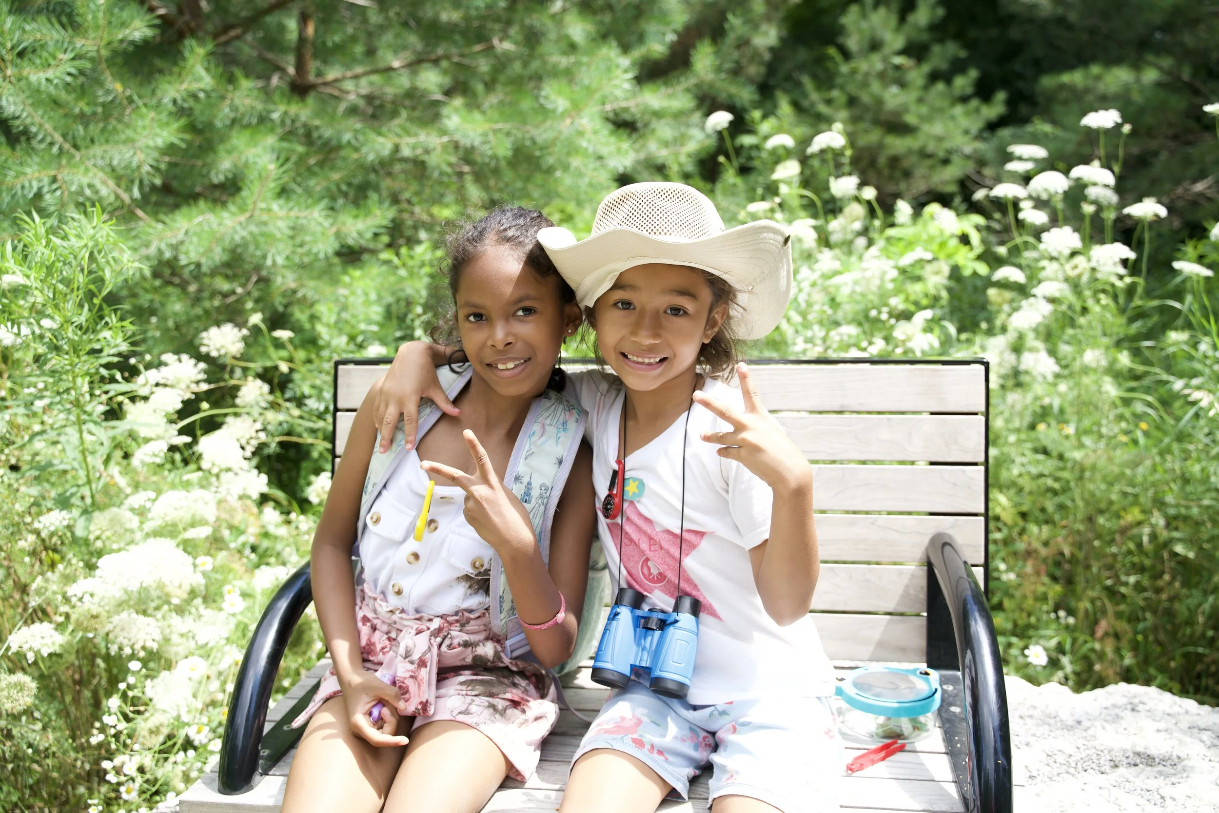 Two young girls sitting on a park bench surrounded by green trees and white flowers, one wearing a large white sunhat and binoculars, and the other wearing a sleeveless top and patterned shorts, smiling and making peace signs.