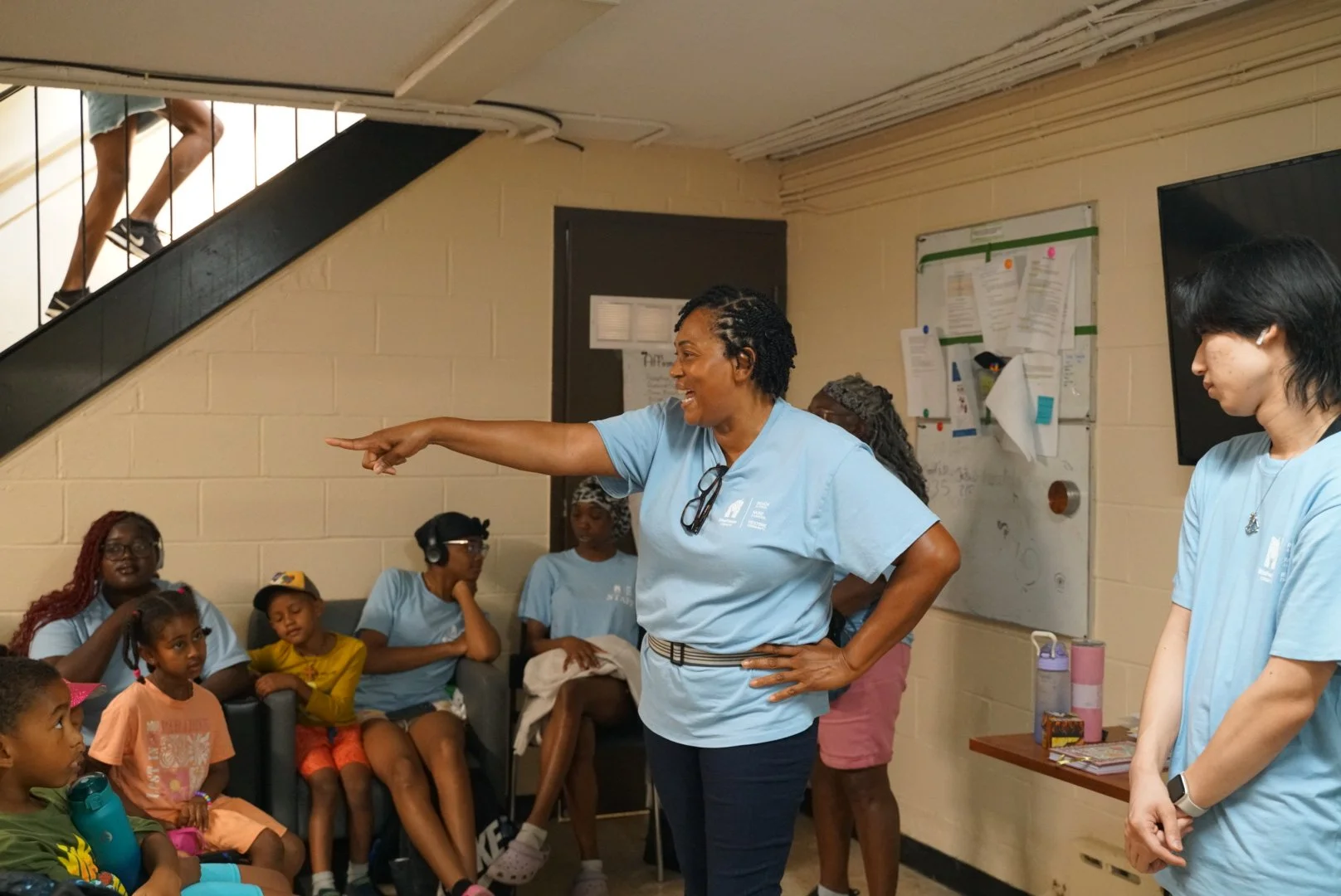 A group of children and women in blue shirts gathered in a room listening to a woman with glasses on her shirt, who is standing and pointing, appearing to be giving a presentation or leading a discussion.
