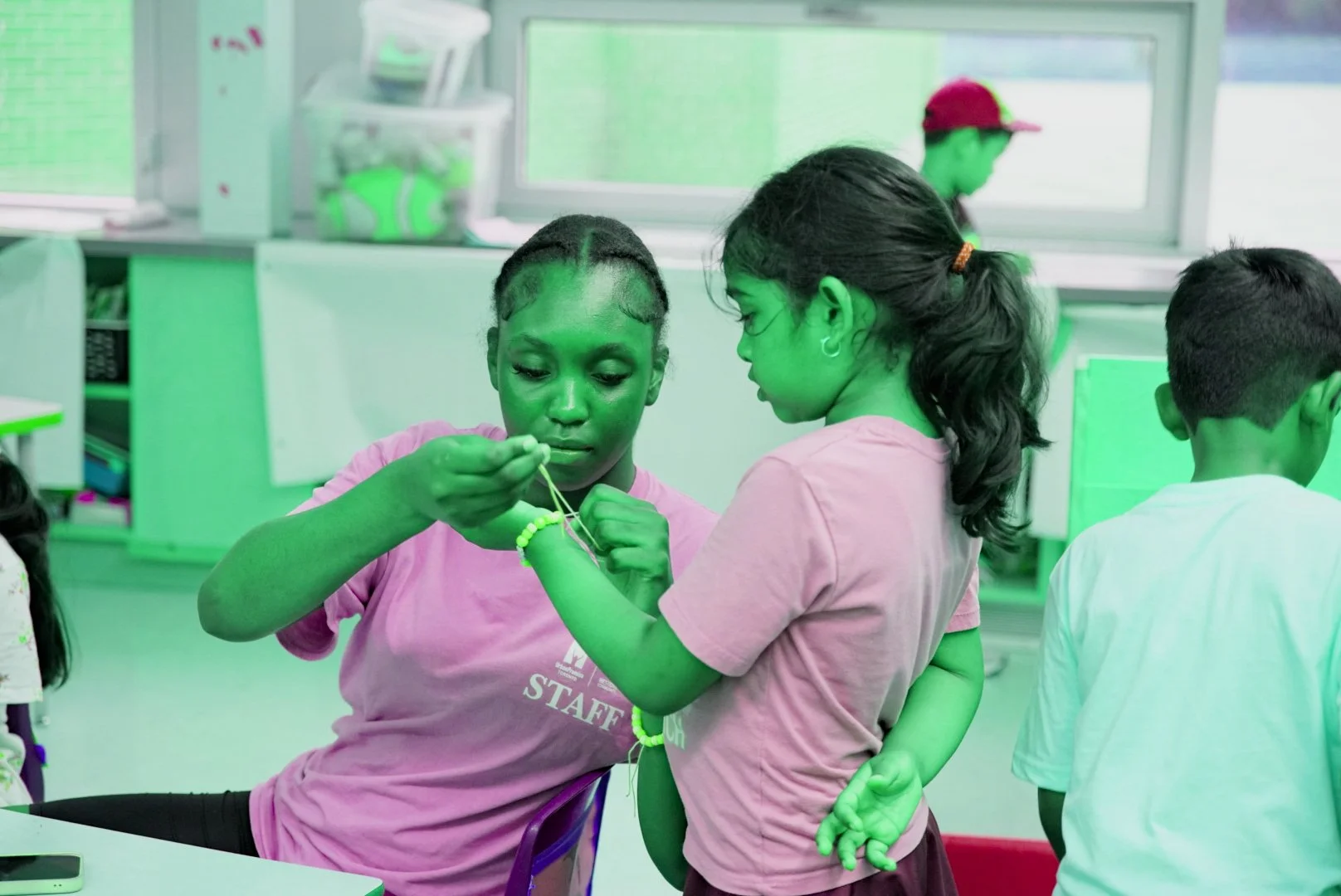 Two girls are helping each other put on a friendship bracelet in a classroom with other children and computers in the background.
