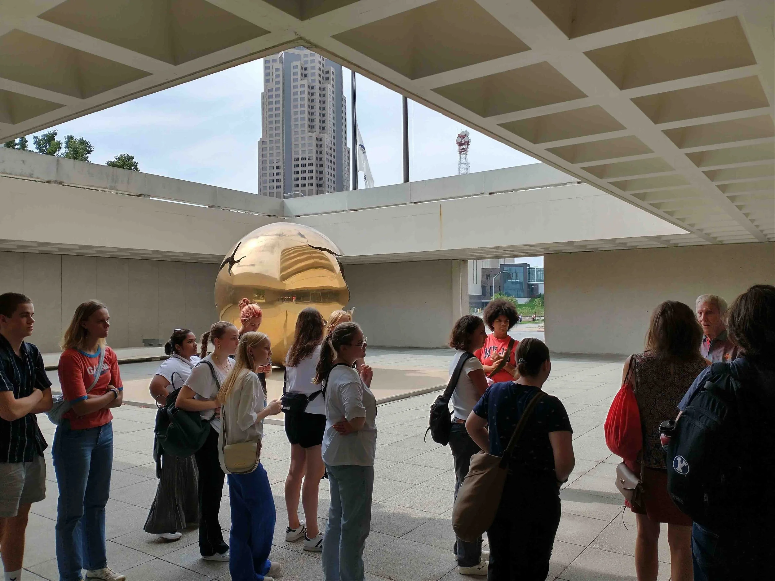 Group of people, mostly young women and girls, standing outdoors under a covered area near a large reflective golden sphere sculpture with a cityscape in the background.