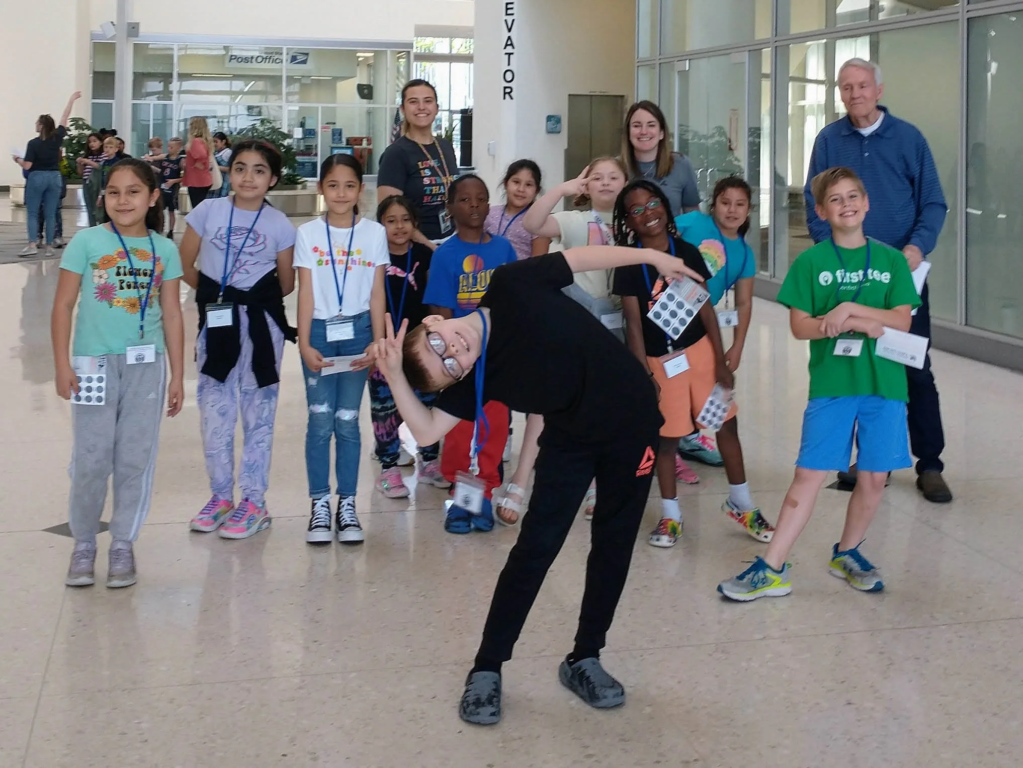 A group of children and two adults standing inside an airport terminal, with some children wearing lanyards and holding papers, and one child in front striking a playful pose.