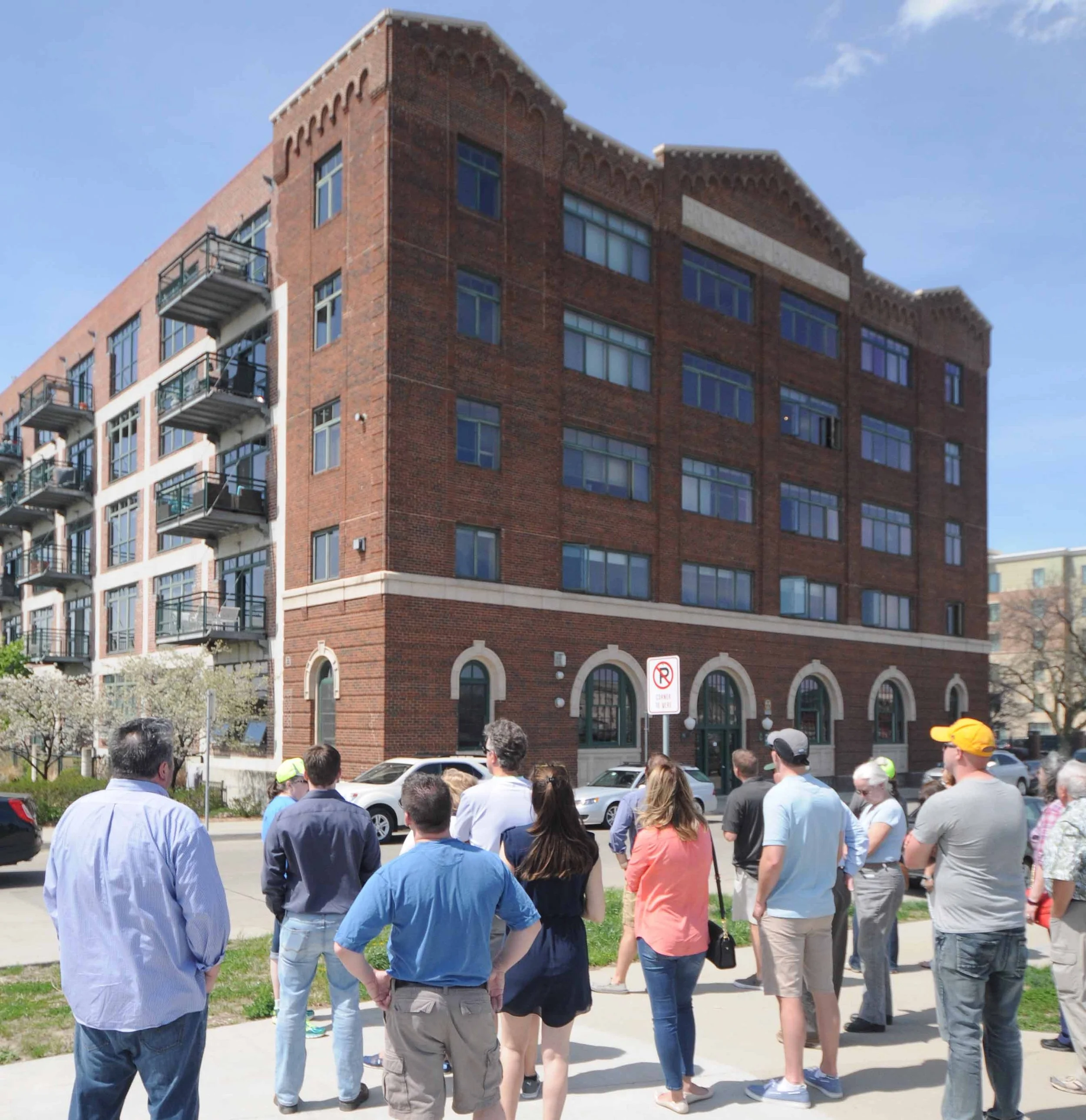 A group of people standing outside on a sidewalk in front of a multi-story brick building with arched windows and balconies, under a blue sky.