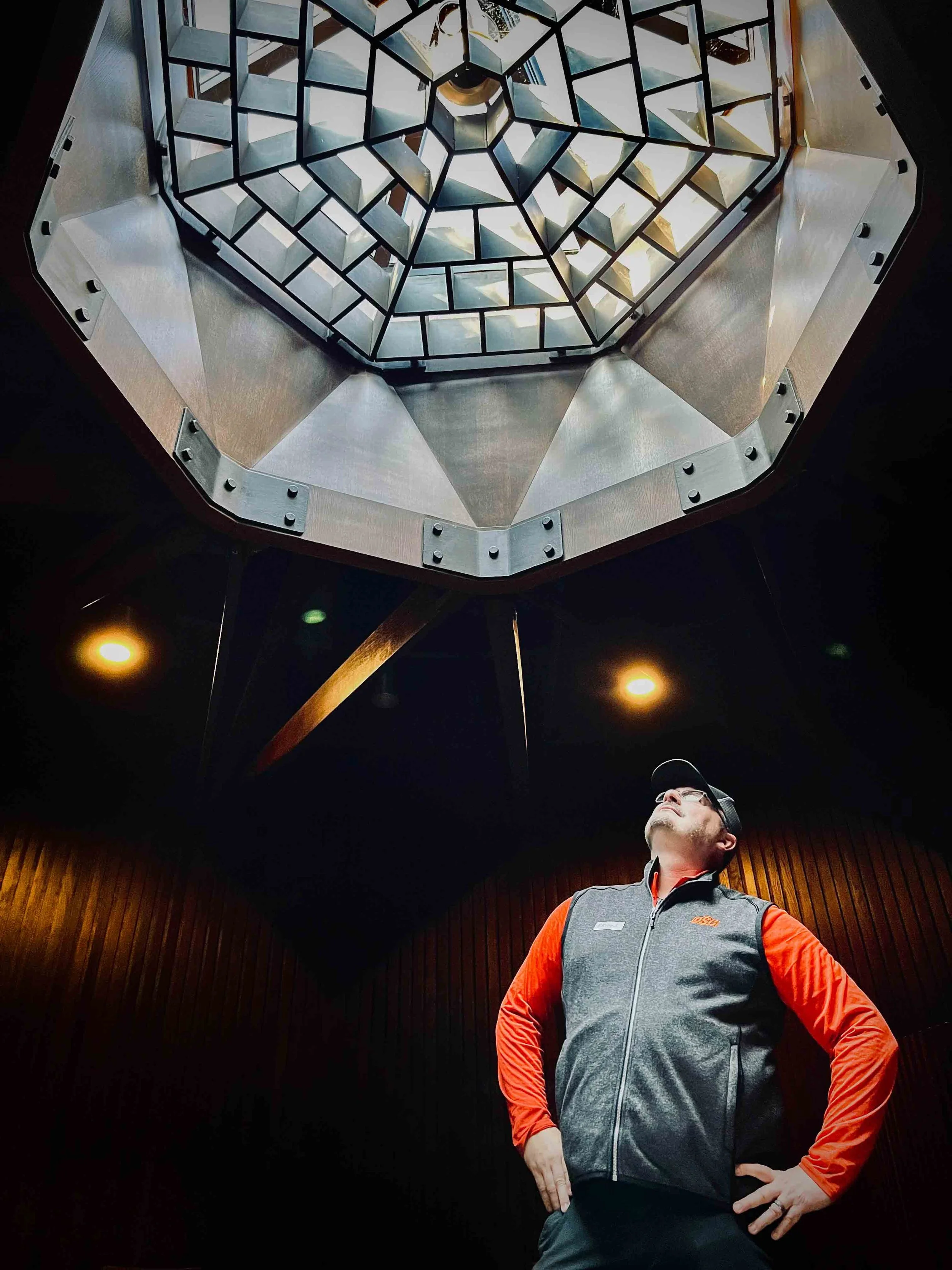A man wearing glasses, a gray vest over an orange shirt, and a cap looks up at an ornate, geometric chandeliers hanging from a dark ceiling in a dimly lit room with wooden walls.