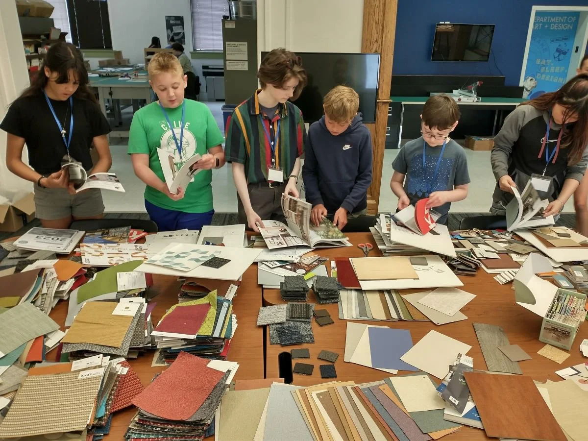Group of children examining a table filled with various sample materials for a design project in a classroom or workshop setting.
