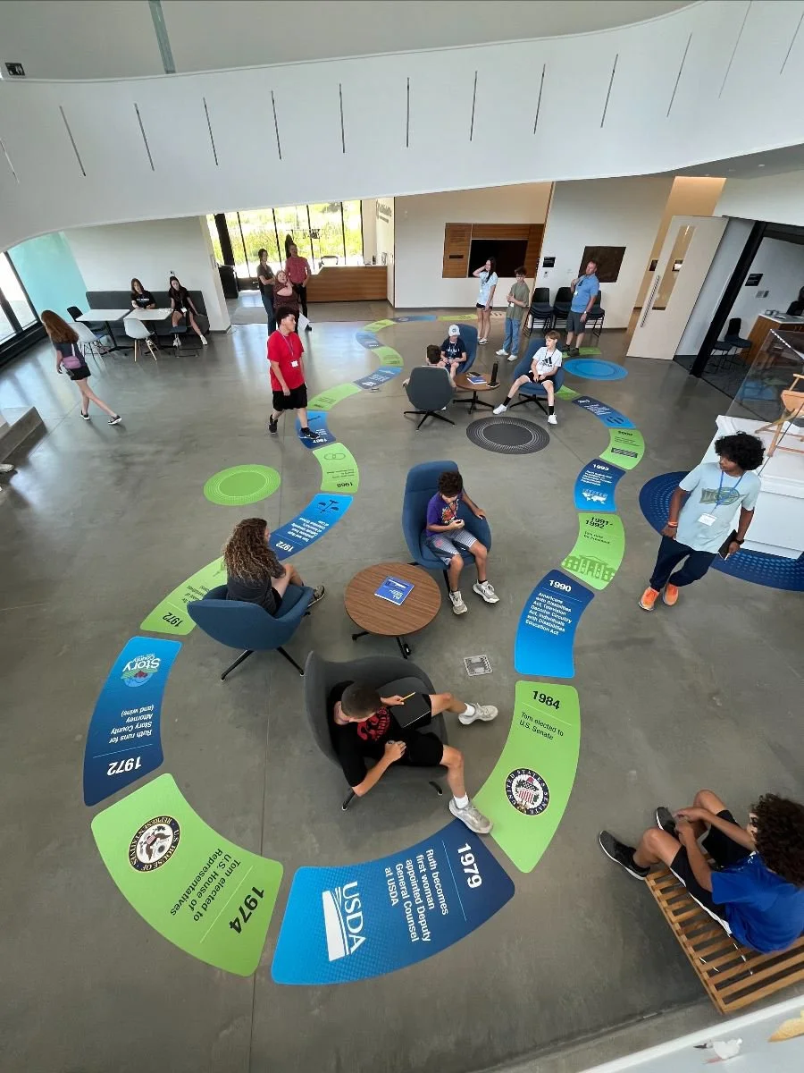 A large indoor space with a spiral timeline on the floor showing historical events from 1941 to 2021. People, including children and adults, are sitting, walking, and standing around the timeline, some using electronic devices and others talking. The room has modern decor with windows and seating areas.