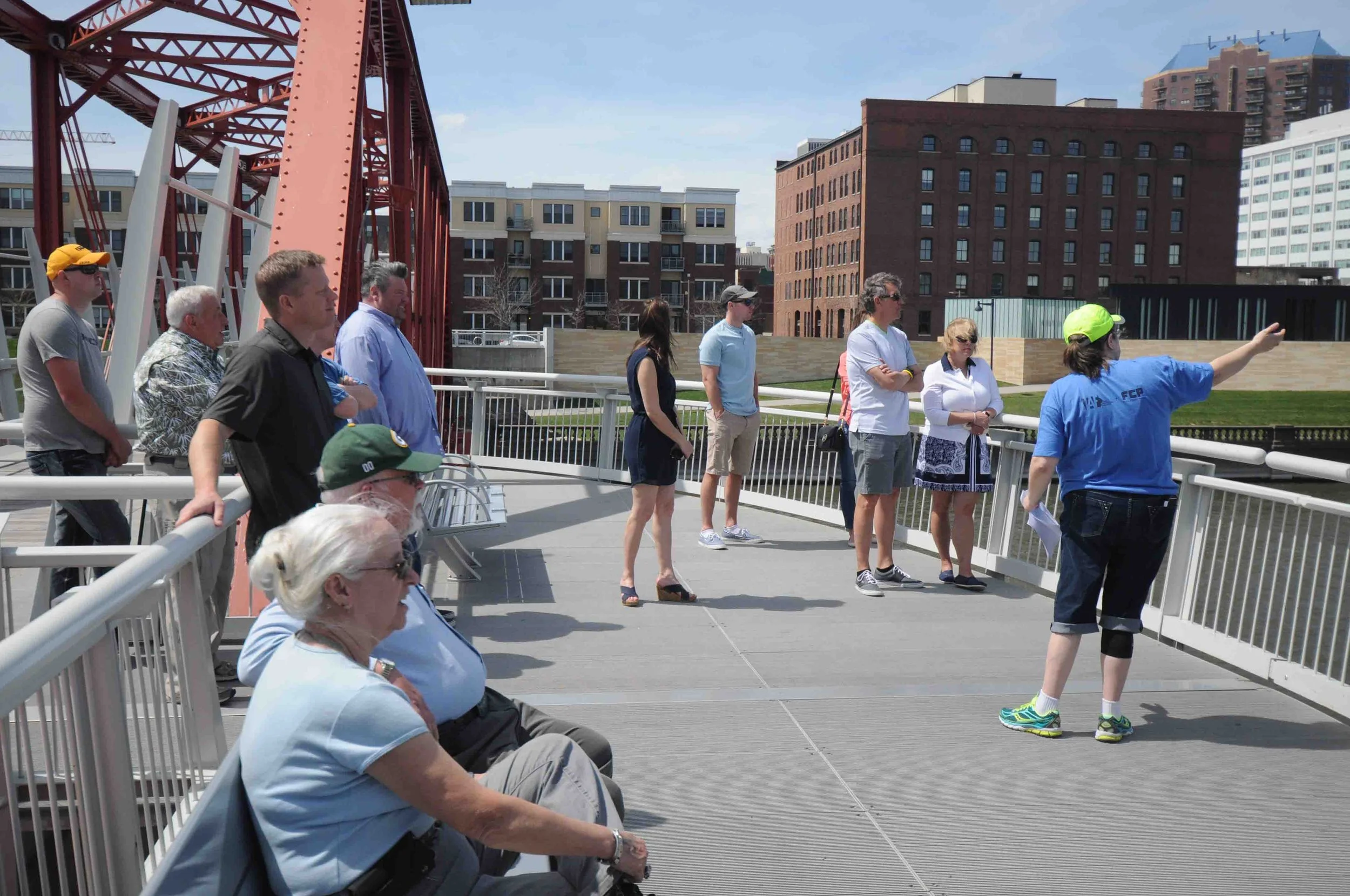 Group of people standing on a bridge or observation deck, looking in one direction while a guide points outward, with urban buildings and a red bridge structure in the background.