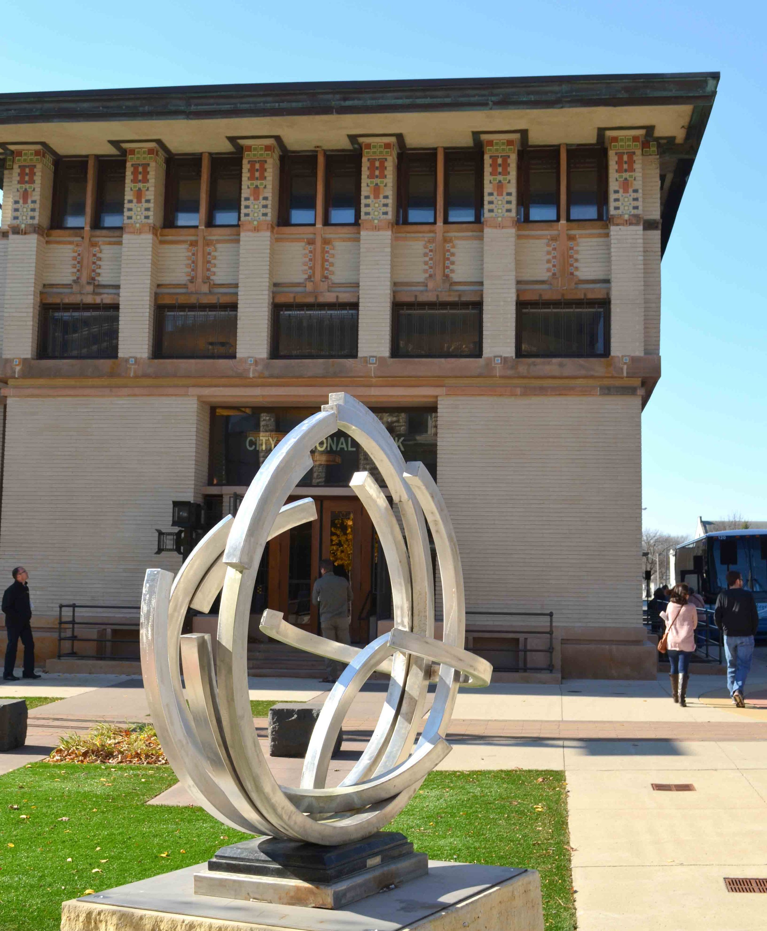 A modern metal abstract sculpture in front of a brick city hall building with people walking by.