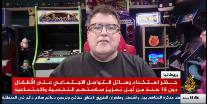 Man speaking in front of a colorful arcade game setup with shelves of toys and gaming memorabilia in the background.