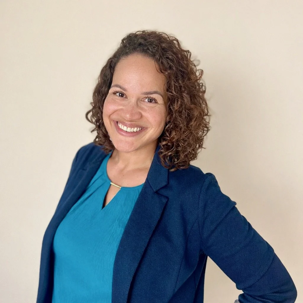 A woman with curly brown hair wearing a blue blazer and a turquoise blouse smiles at the camera, standing against a plain light-colored background.