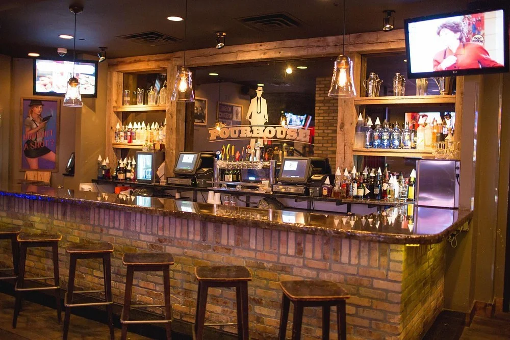 A bar with a brick front, topped with a polished countertop, and four wooden bar stools. Behind the bar are shelves with bottles and glasses, illuminated by warm lighting. Two mounted TV screens are visible, one showing a woman with curly hair and a red top, and the other with a menu. The bar has a sign that reads "The Pourhouse," and a framed vintage poster on the wall.