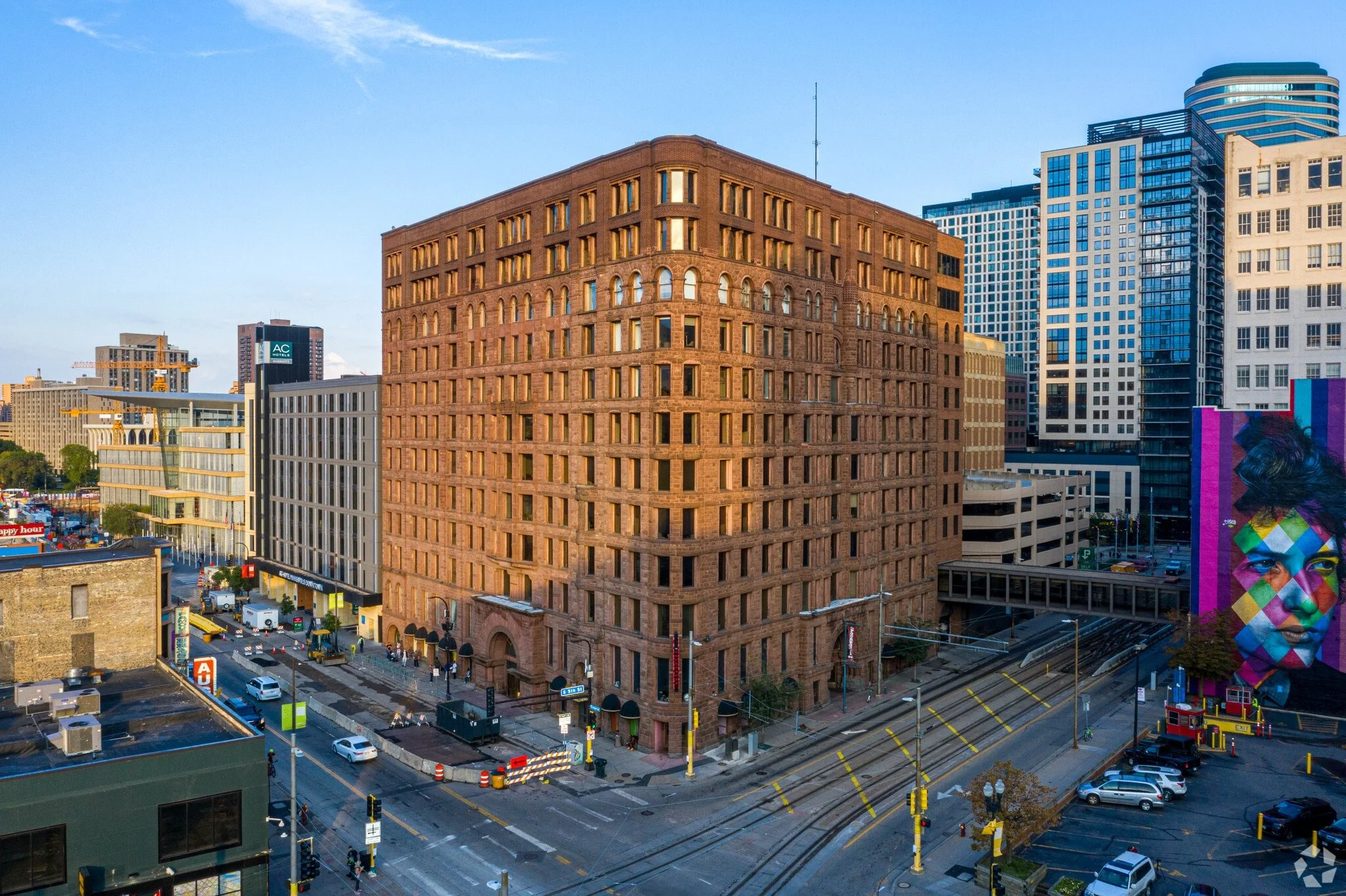 A cityscape featuring a large, historic brick building at the corner of the street, surrounded by modern skyscrapers. The street below has moving cars, parked vehicles, and trolley tracks, with some street signs and traffic signals visible. Lumber Ex
