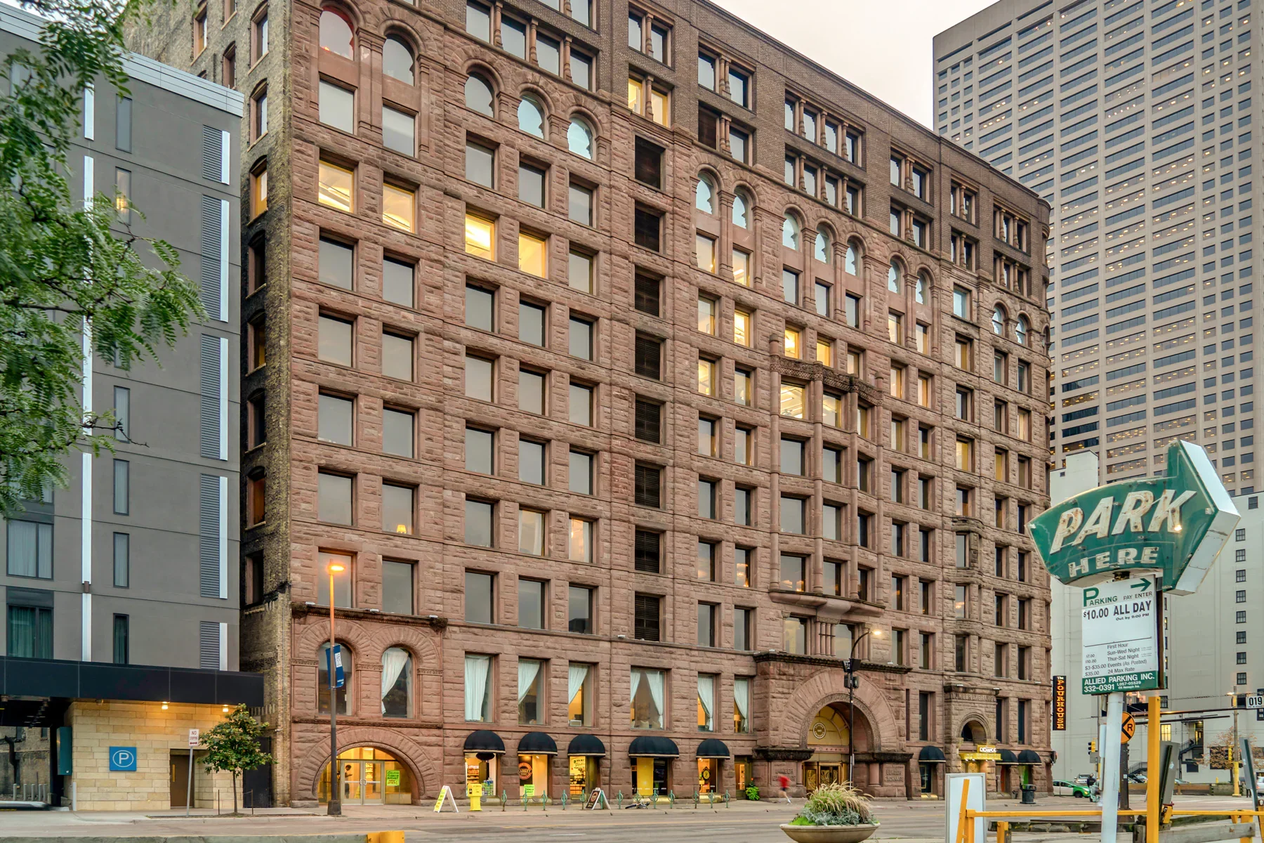 A tall historic brick building with multiple lit windows, located in an urban area with modern buildings nearby. There is a parking sign on the street. Lumber Exchange Building in Minneapolis, Minnesota