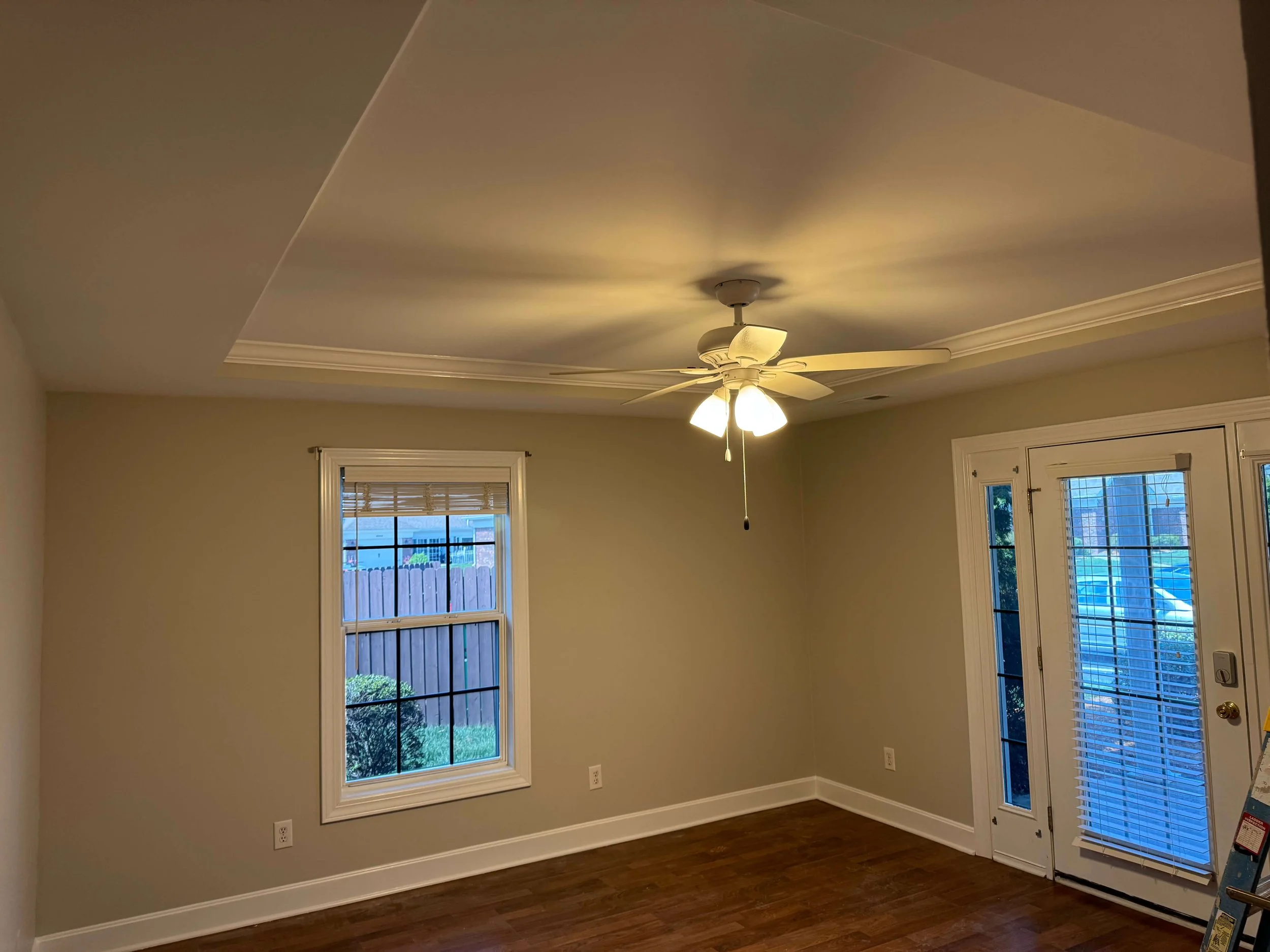 Empty room with beige walls, hardwood floors, three windows with blinds, a white ceiling fan with lights, and a door with a screen.