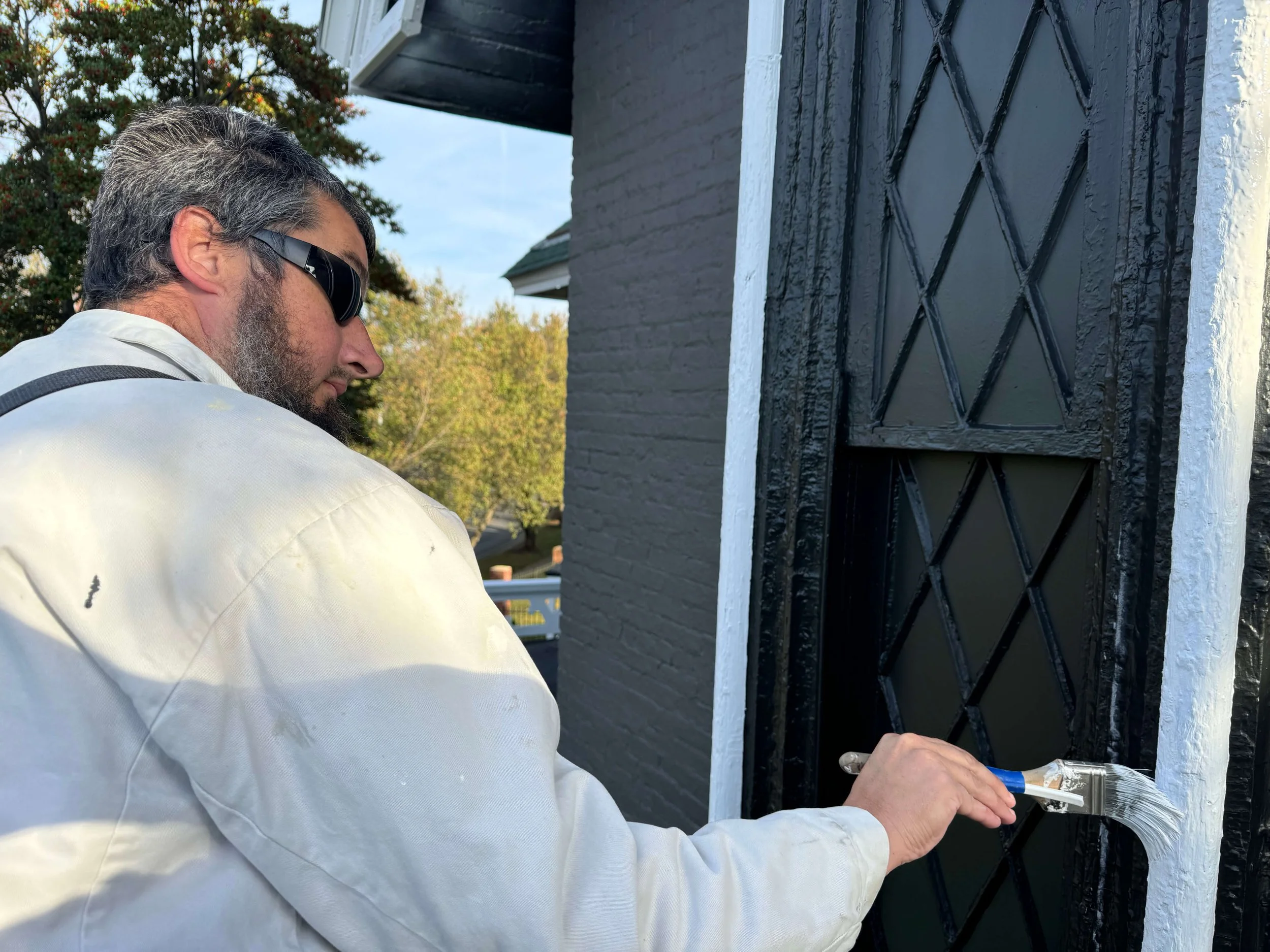 A man wearing sunglasses and a white shirt is working on a black door with a diamond lattice design, sealing or painting the door on a house exterior.