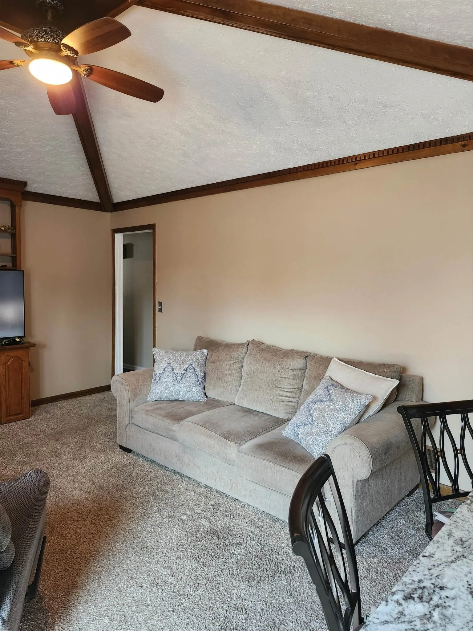 Living room with beige couch, patterned pillows, ceiling fan, and a corner TV stand with a television. Carpeted floor and a doorway leading to another room.