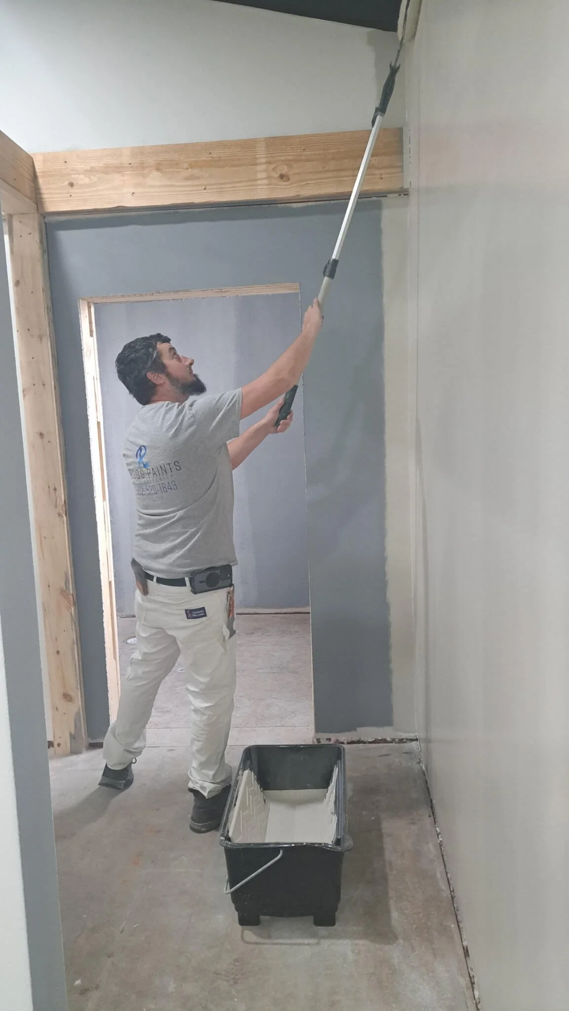 Man painting a wall with a roller using white paint while standing on the floor in a room under construction.