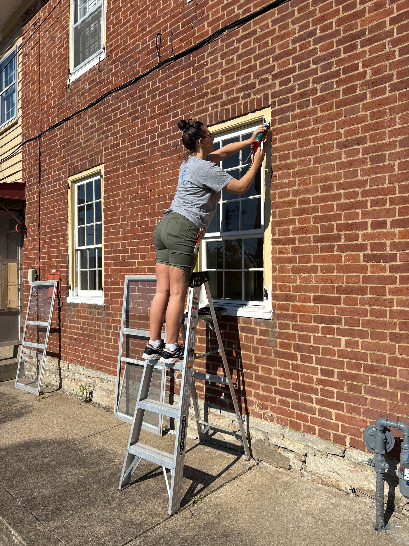 A woman standing on a step ladder cleaning a window on a brick building, using a cleaning tool with a long handle. There are additional ladders and a water meter visible outside on a sidewalk.