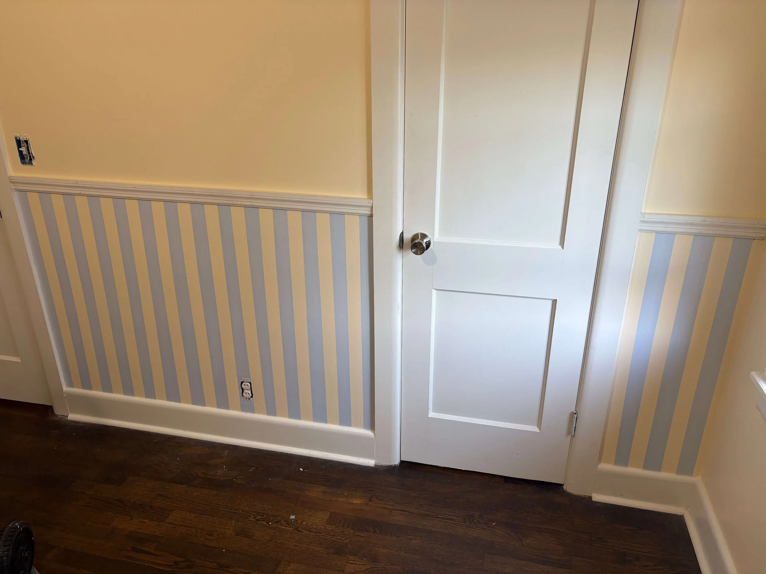 A residential room corner with a closed white door featuring a round doorknob, striped wallpaper in light blue and beige, a yellow painted wall, and dark hardwood flooring. An electrical outlet and a cable socket are visible on the wall.