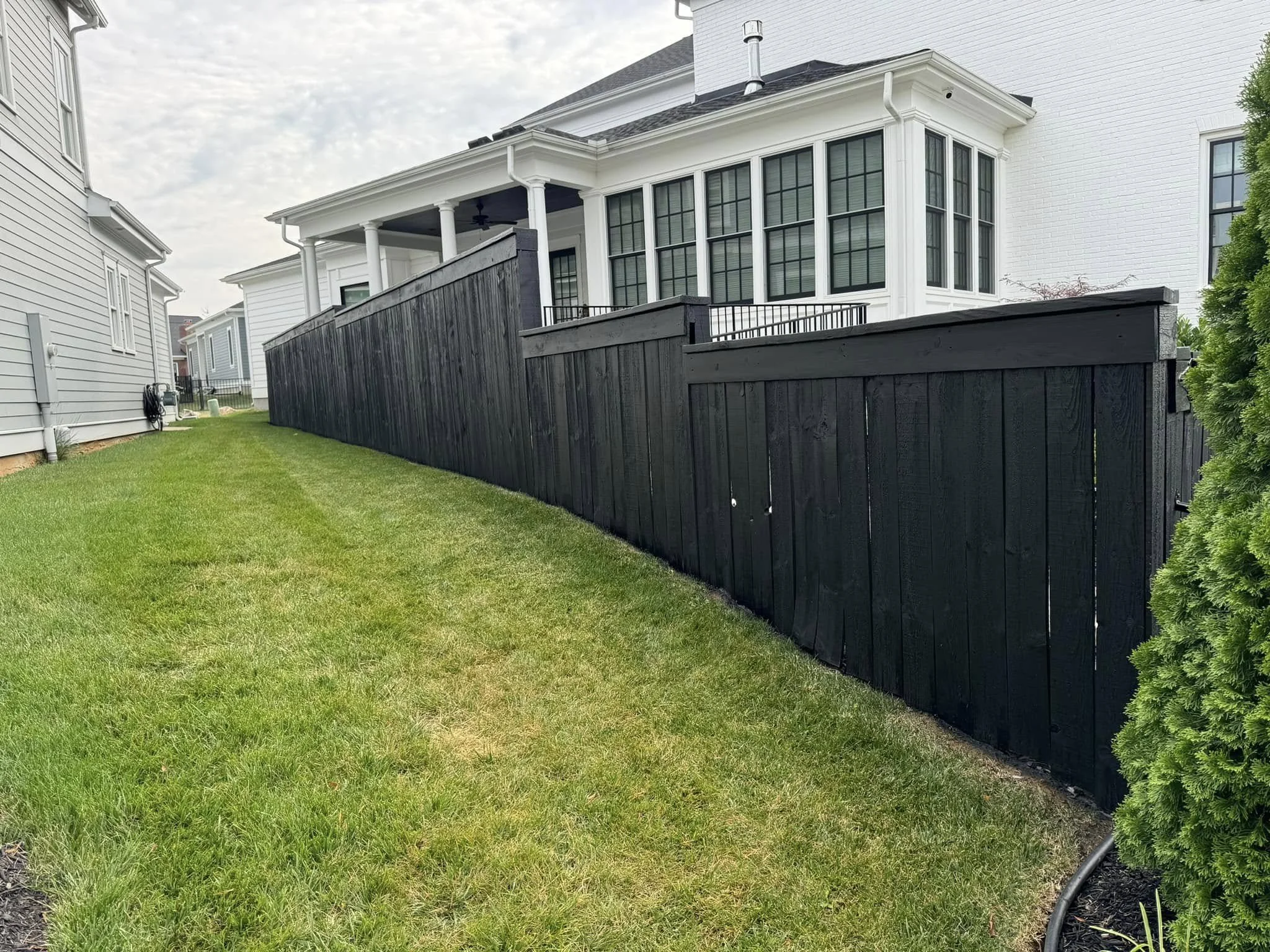 Backyard with a green lawn, black wooden fence, white house with large windows and porch, and neighboring houses.