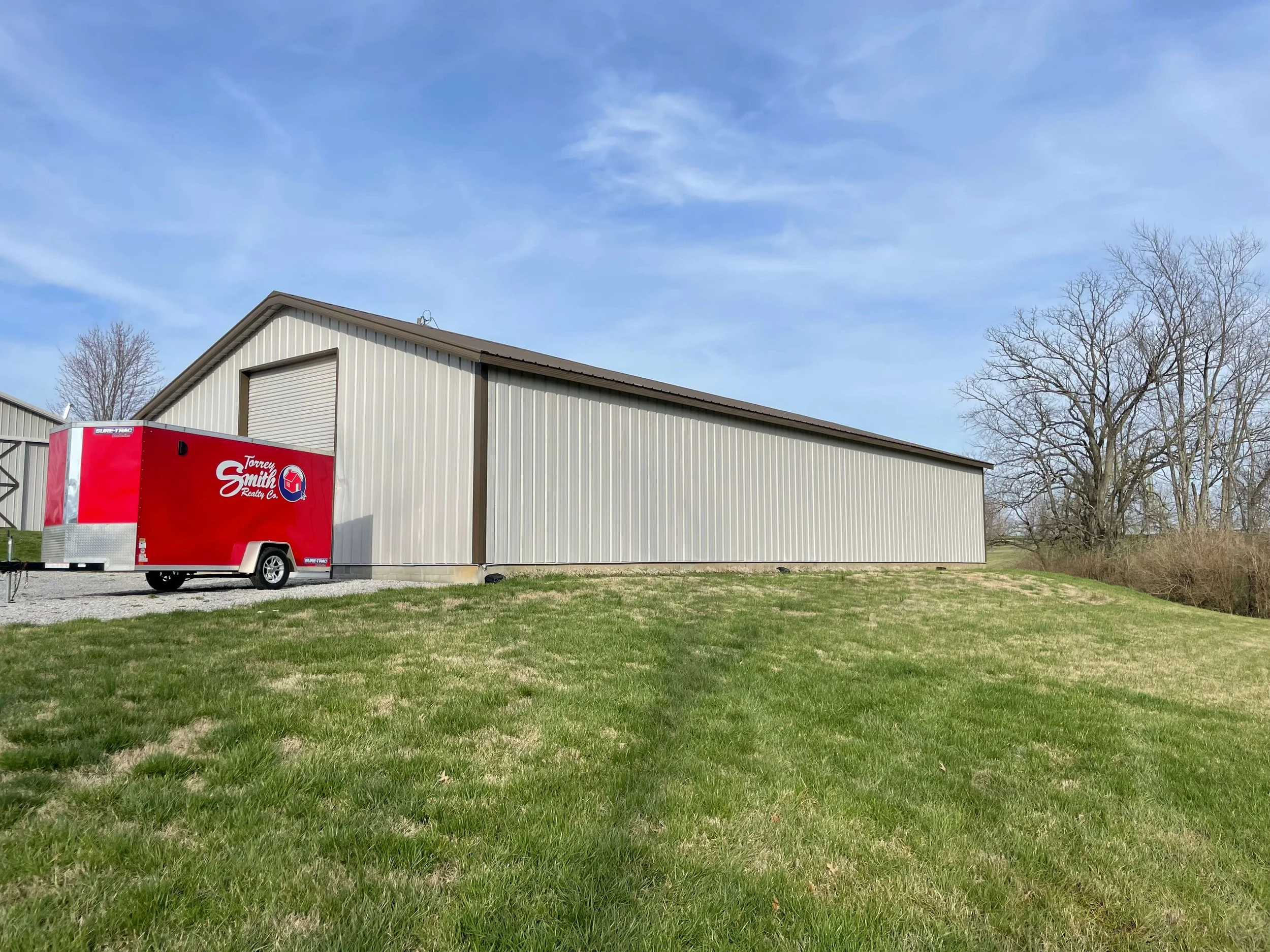 A large metal building with a sloped roof and a roller door, situated behind a grassy area with some trees nearby. Part of a red and white trailer from Torrey Smith Realty Co. is visible in the foreground.
