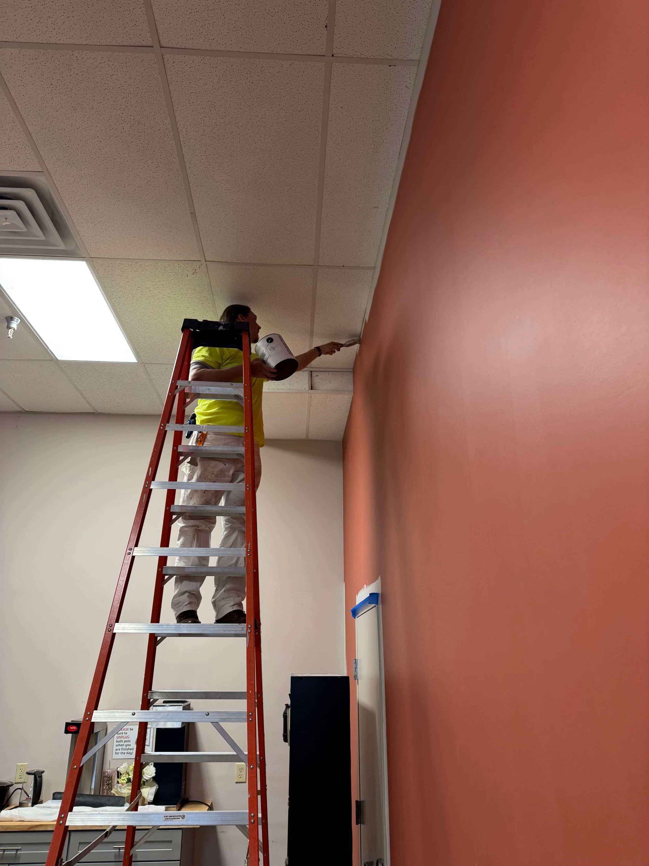 A person standing on a red ladder painting a ceiling with a paint roller, in a room with a pink wall and beige ceiling tiles.