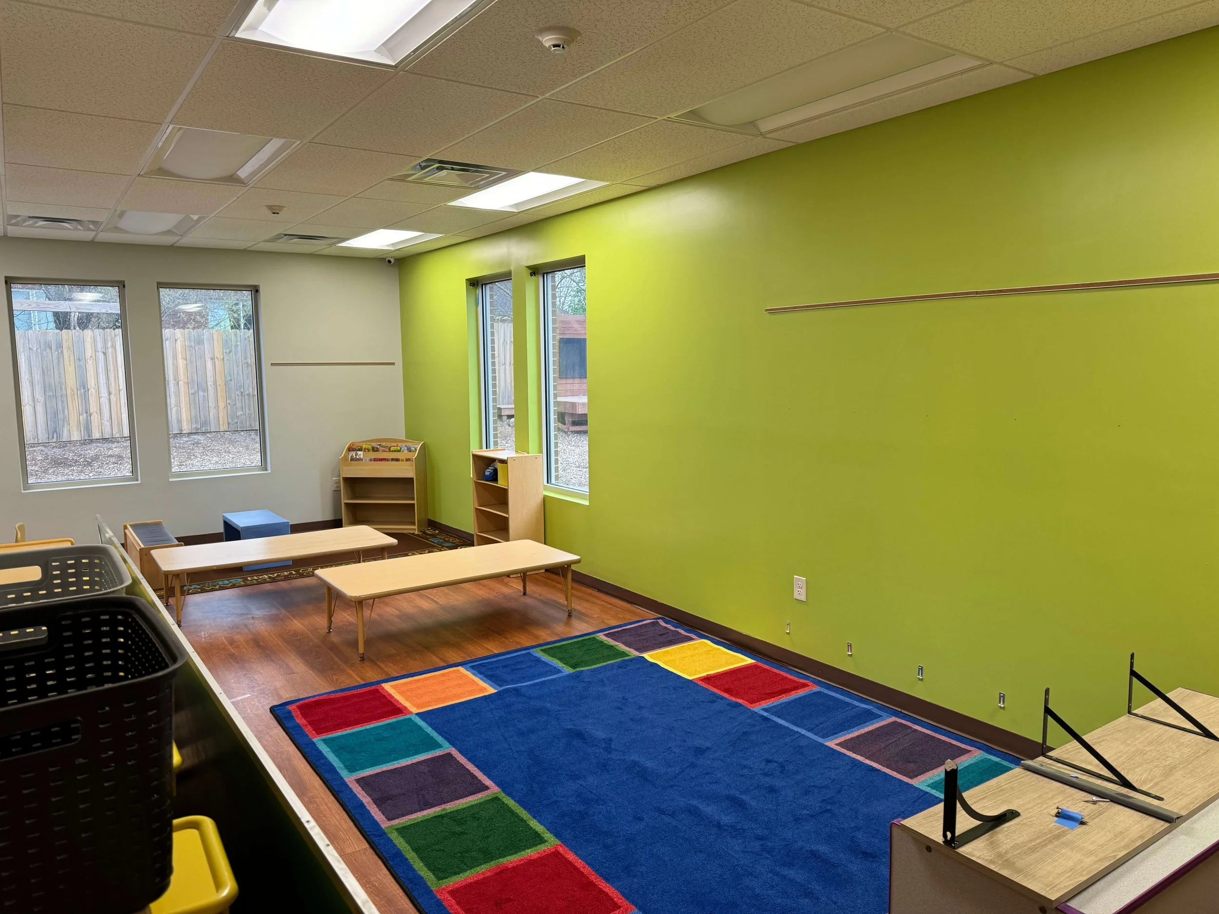An empty classroom with bright green and light beige walls, large windows, a colorful rug, and small furniture.