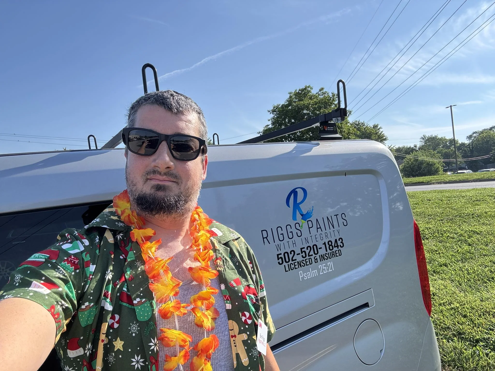 A man with short gray hair and a beard, wearing sunglasses, a colorful Hawaiian shirt, and an orange lei, taking a selfie in front of a white service vehicle with company information, outdoors on a sunny day with blue sky and trees in the background.
