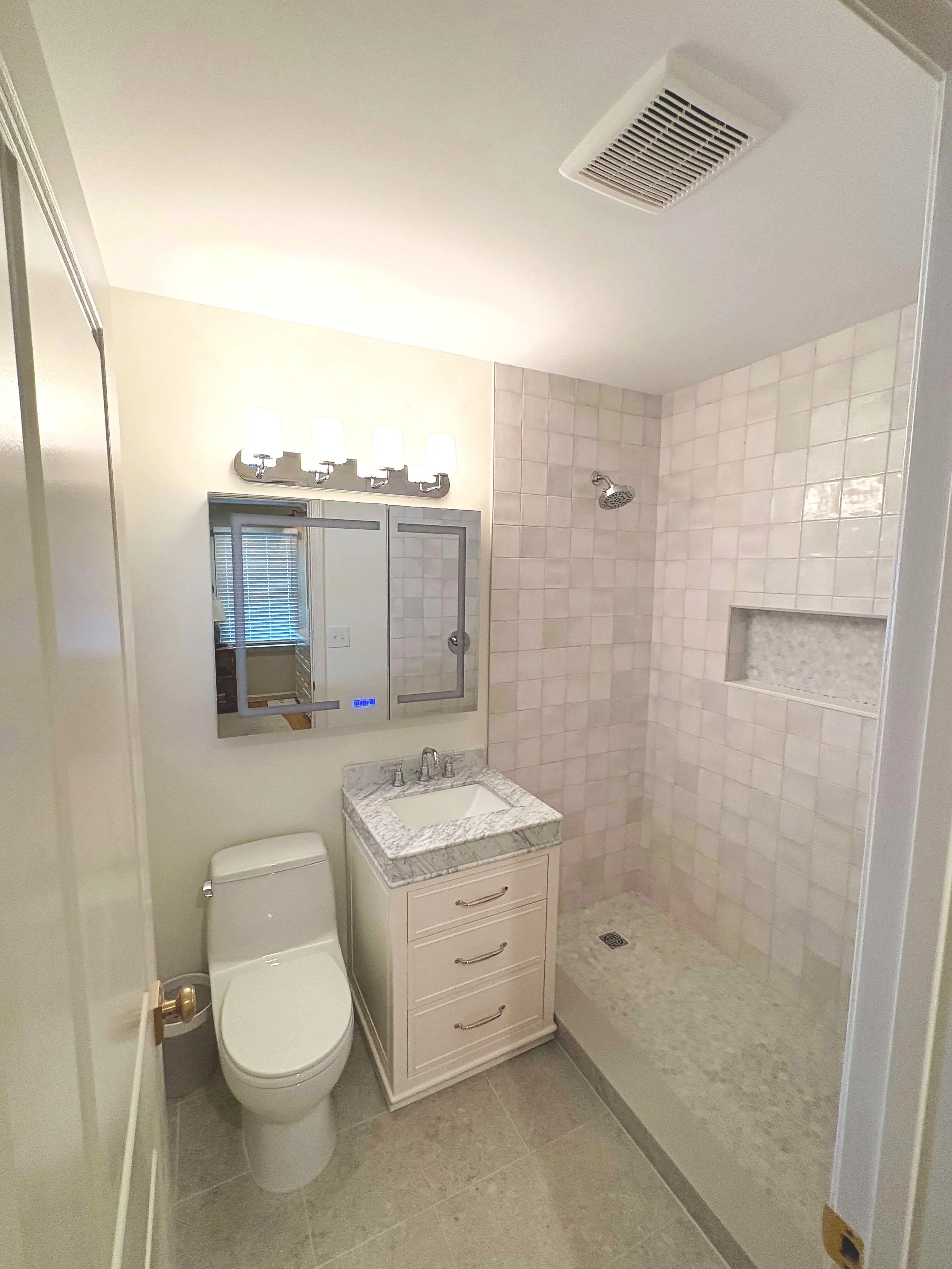 Bathroom with a toilet, a small vanity with a sink and mirror, a shower area with a tiled wall, and a vent on the ceiling.