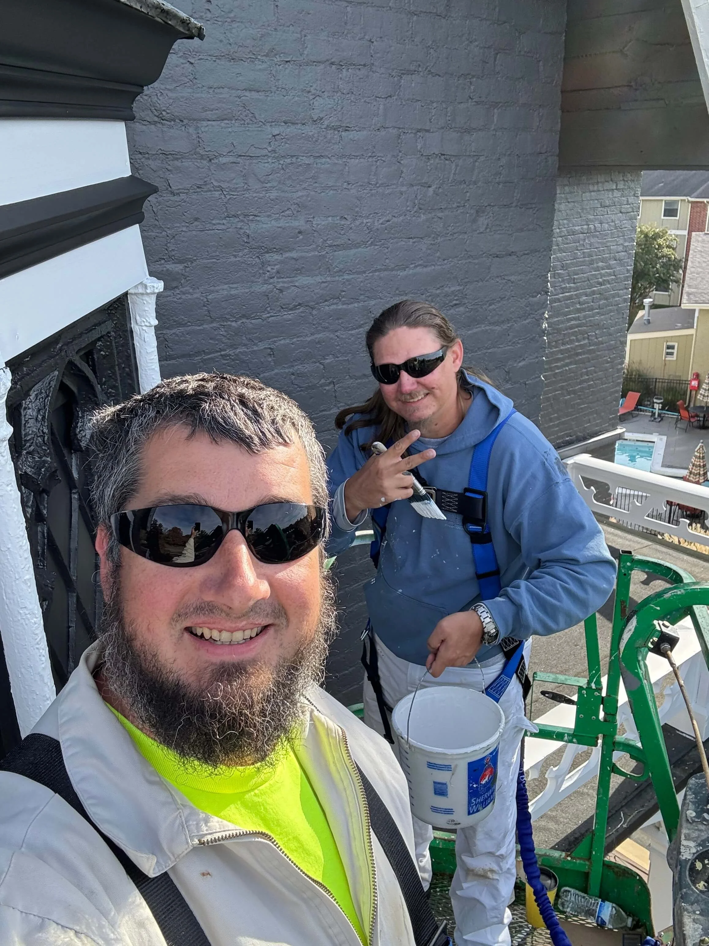 Two men, one with gray hair and beard wearing sunglasses and a reflective vest, and the other with long brown hair and sunglasses, smiling and pointing, on a rooftop with clear skies, brick walls, and outdoor furniture in the background.