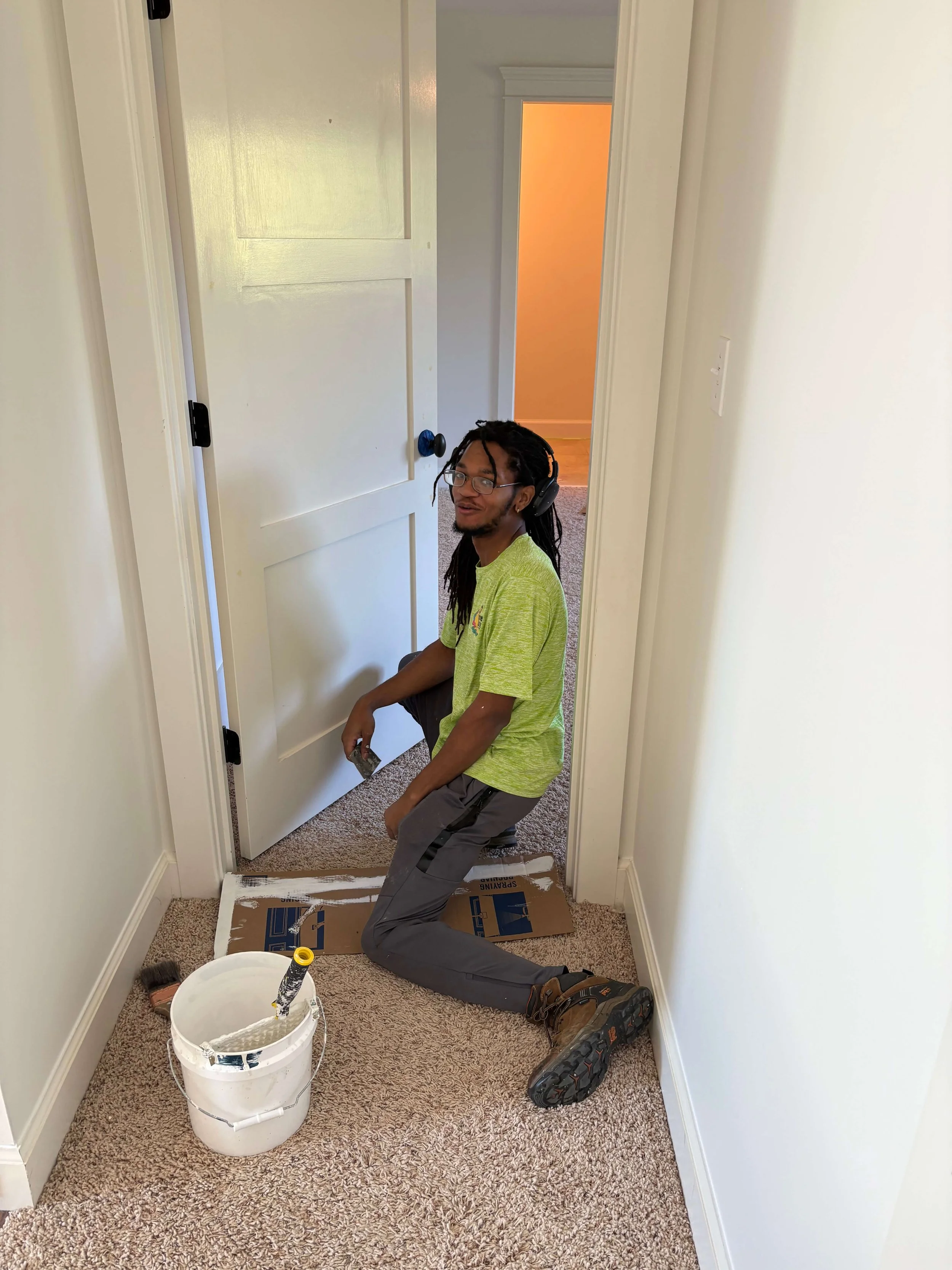 A man kneeling on a carpeted hallway floor, painting the baseboard, with painting supplies including a bucket of paint, roller, and brush nearby, and a cardboard cover on the floor, wearing headphones, glasses, and a green shirt.