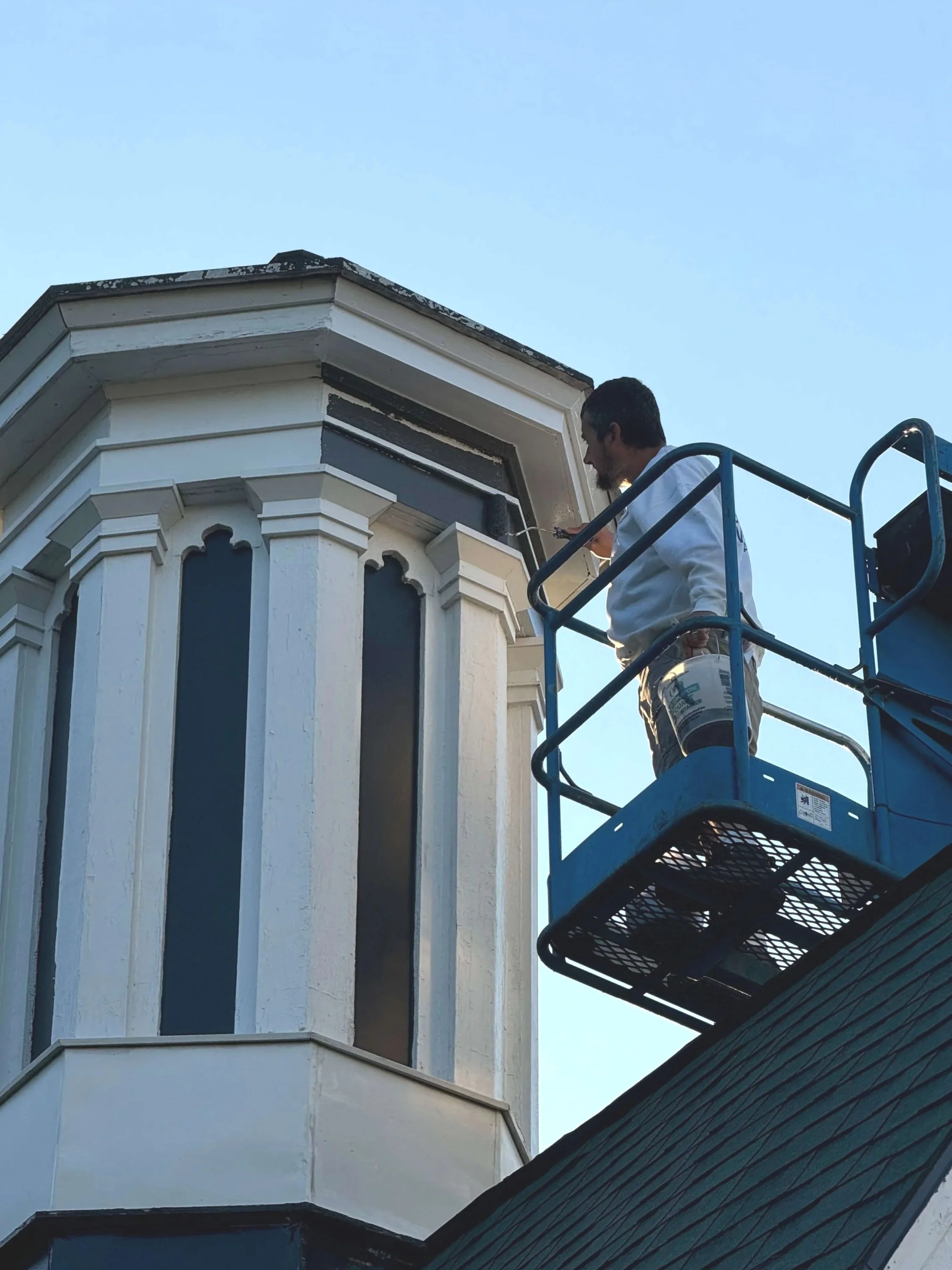 A worker on a lift painting the exterior trim of a Victorian-style house, focusing on the window and decorative molding.
