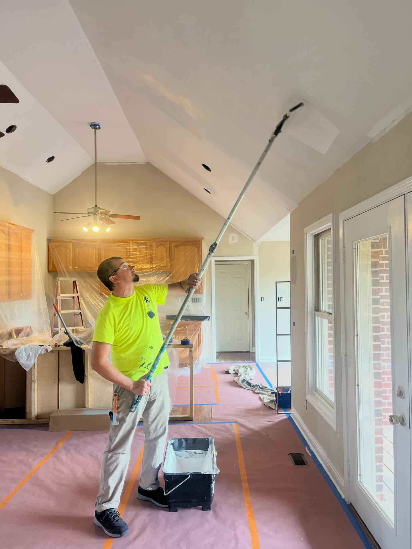 A man in a bright yellow shirt and white pants painting a ceiling with a long roller brush. The room has large windows, a ceiling fan, and cabinets covered with plastic for painting. The floor is protected with cardboard and painter's tape.
