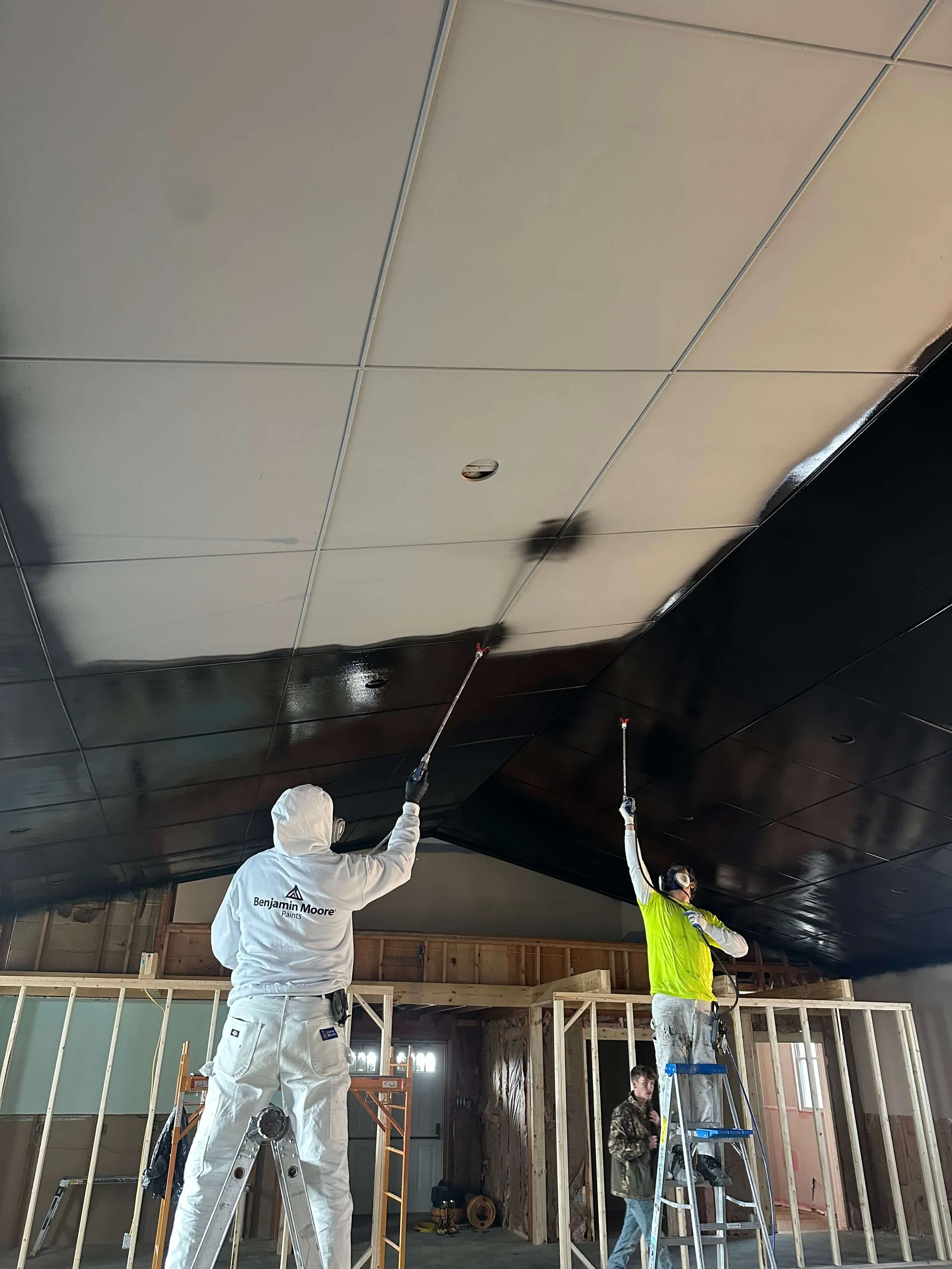 Construction workers installing a ceiling, one is wearing a white coveralls with 'Benjamin Moore Paints' logo and two others are on ladders working on the ceiling.