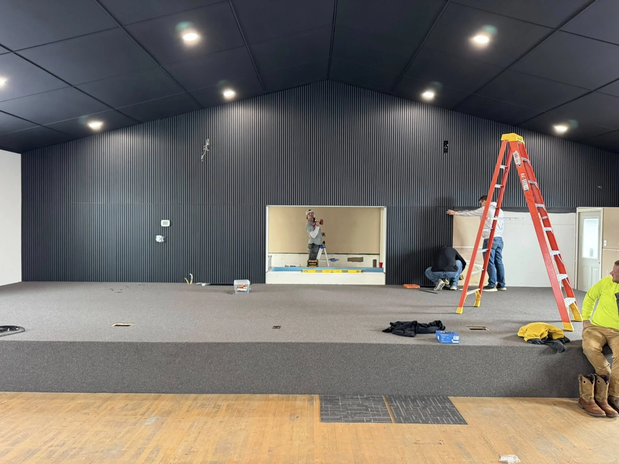 Construction workers installing black vertical paneling on a high wall inside a building. There is a red ladder, tools, and equipment visible, with workers working on the wall and ceiling installation.
