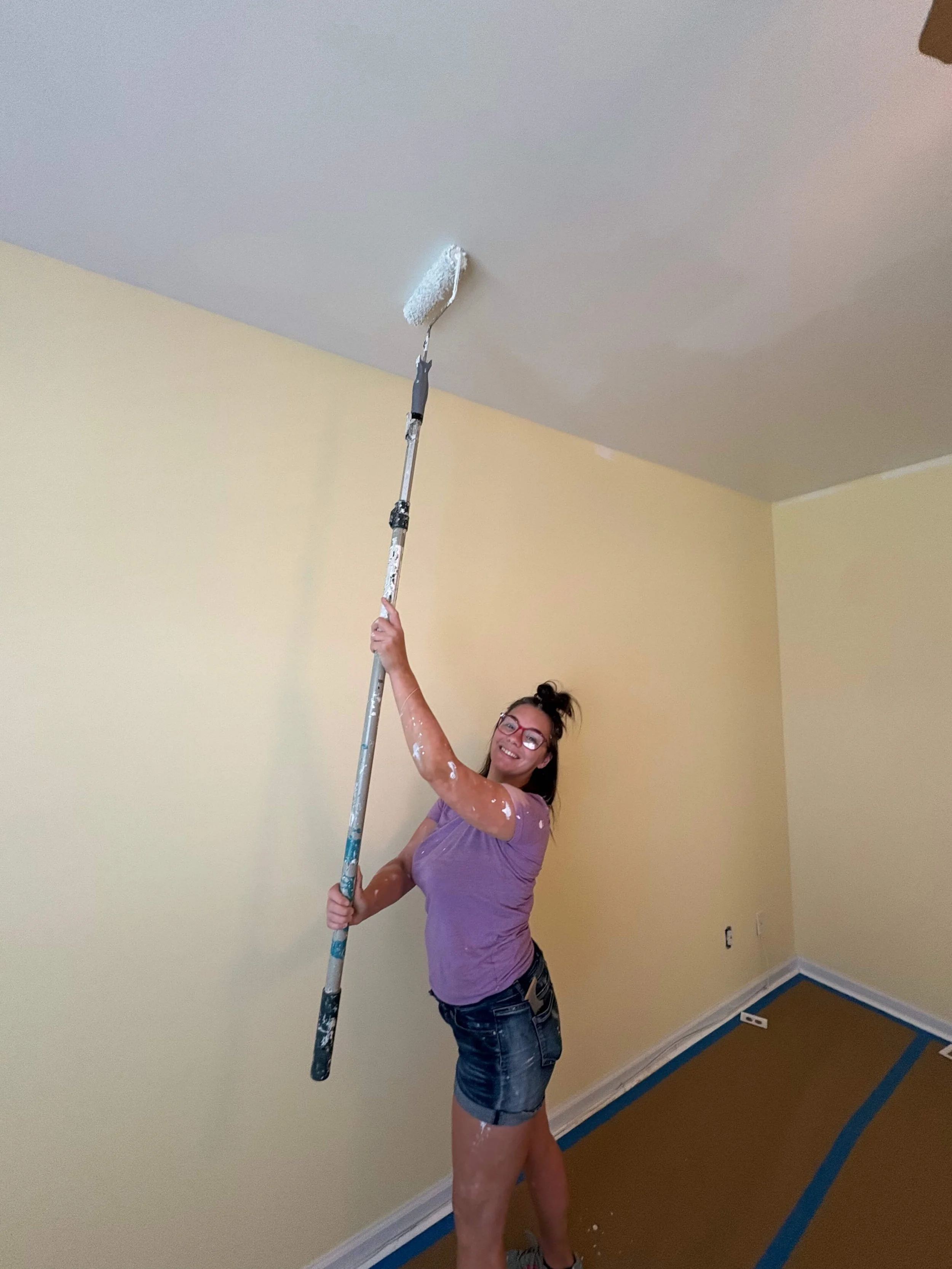 Woman with glasses and a purple t-shirt painting a ceiling with a roller brush, standing in a room with yellow walls and blue painter's tape along the baseboards.