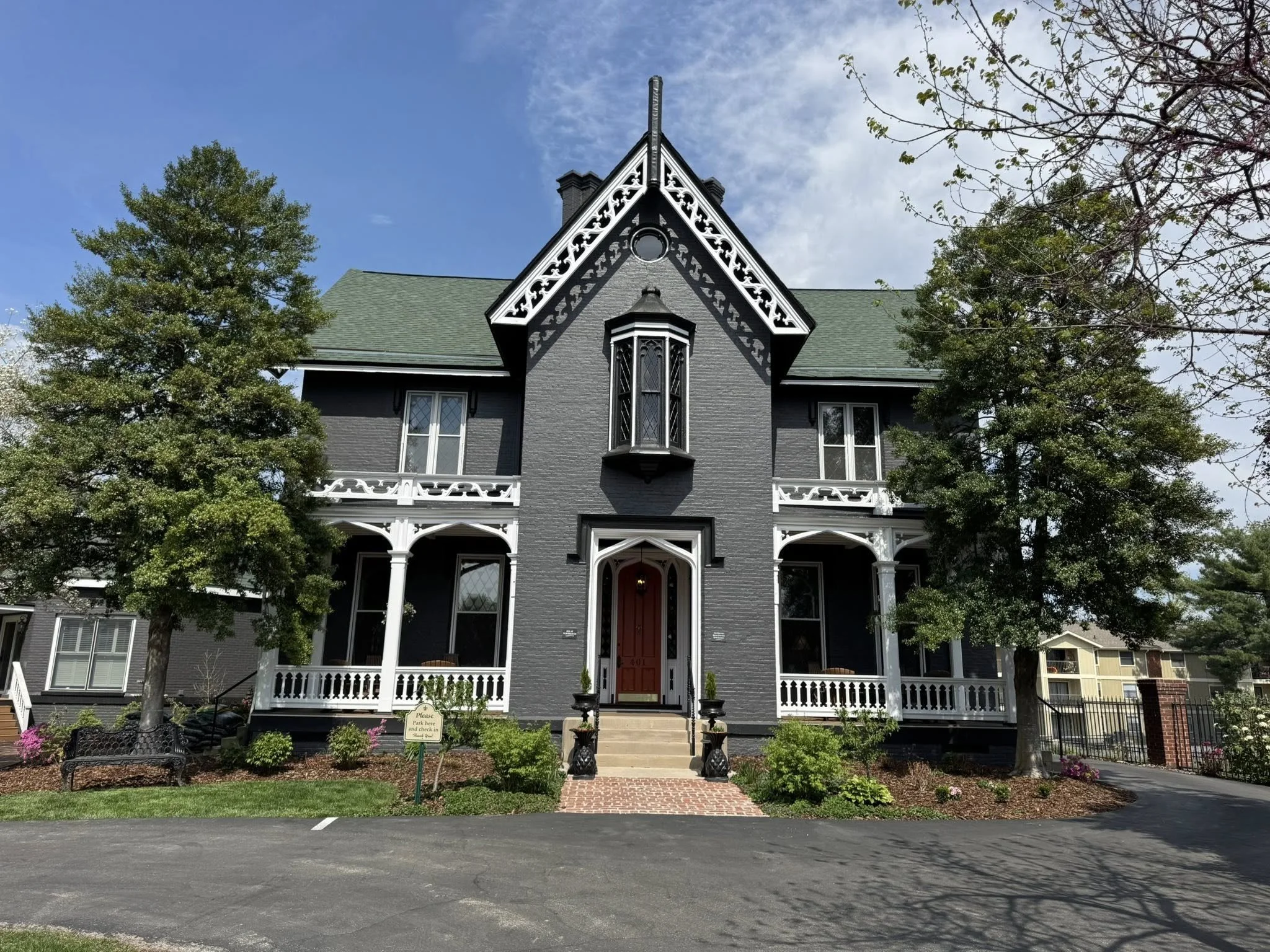 A two-story Victorian-style house painted dark gray with white decorative trim. It has a bay window above the front door and a steeply pitched roof. The front yard has a small garden with flowers, a bench, and a sign that reads 'Please park here and check in.' There are trees on either side of the house, and the driveway and street are visible in the foreground.