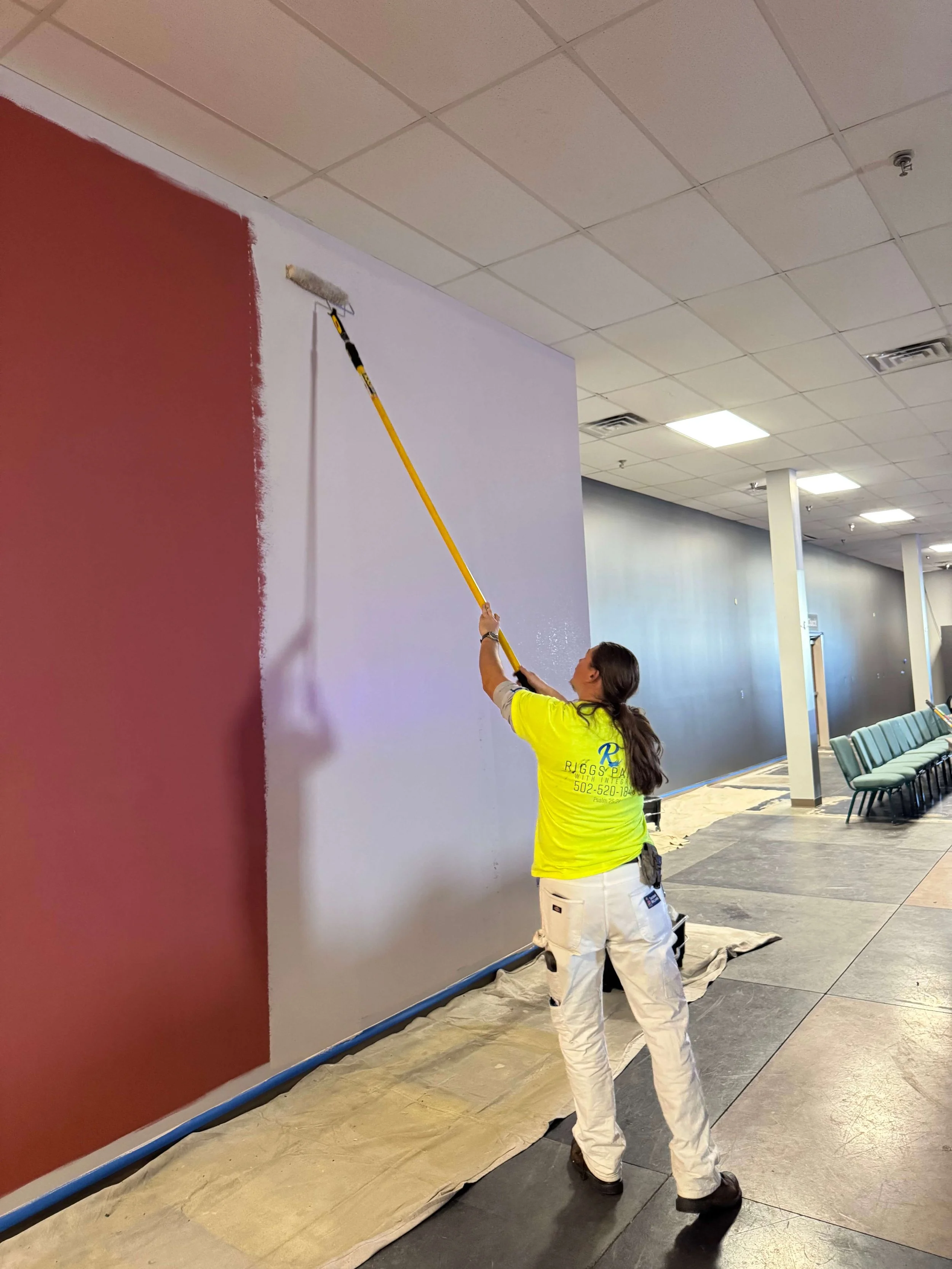 A person painting a wall in a public space, using a roller brush with a long handle. They are wearing a bright yellow shirt, white pants, and black shoes. The wall is partly painted red, and a drop cloth covers the floor beneath the area being painted.