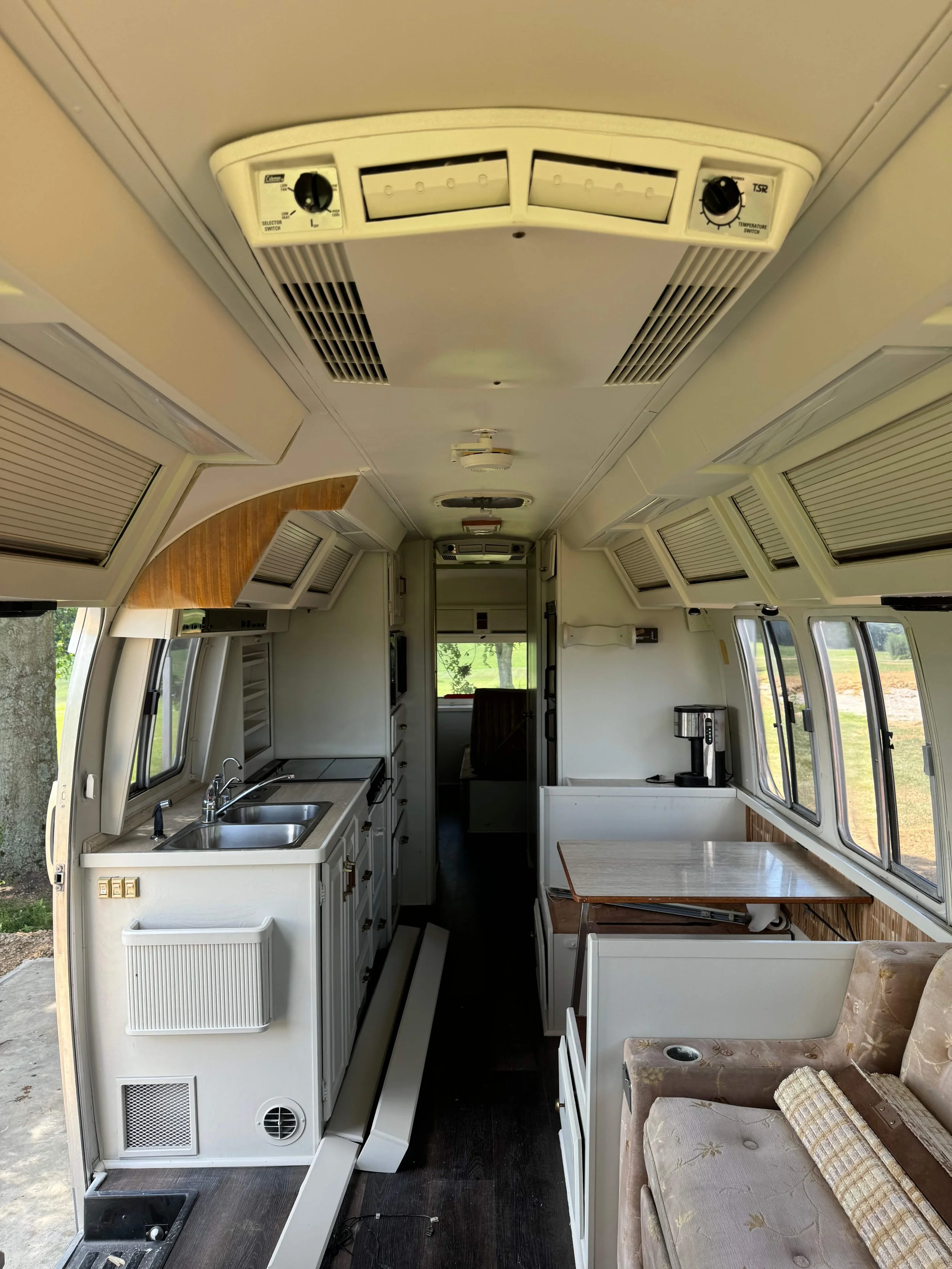 Interior of a converted bus with a small kitchen area including a sink, countertop, and cabinets, as well as windows, a seating area with a couch and pillows, and various ventilation and air conditioning units on the ceiling.