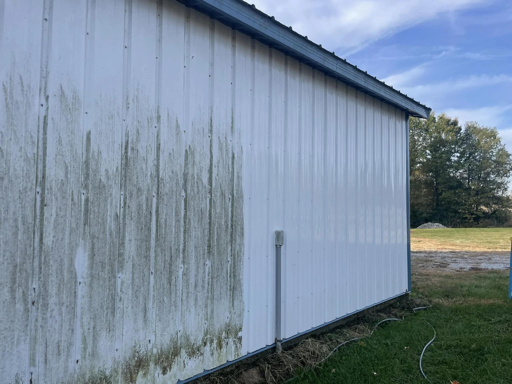 The side of a large building with metal siding, some moss or algae growth, and a metal roof, with trees and a partly cloudy sky in the background.