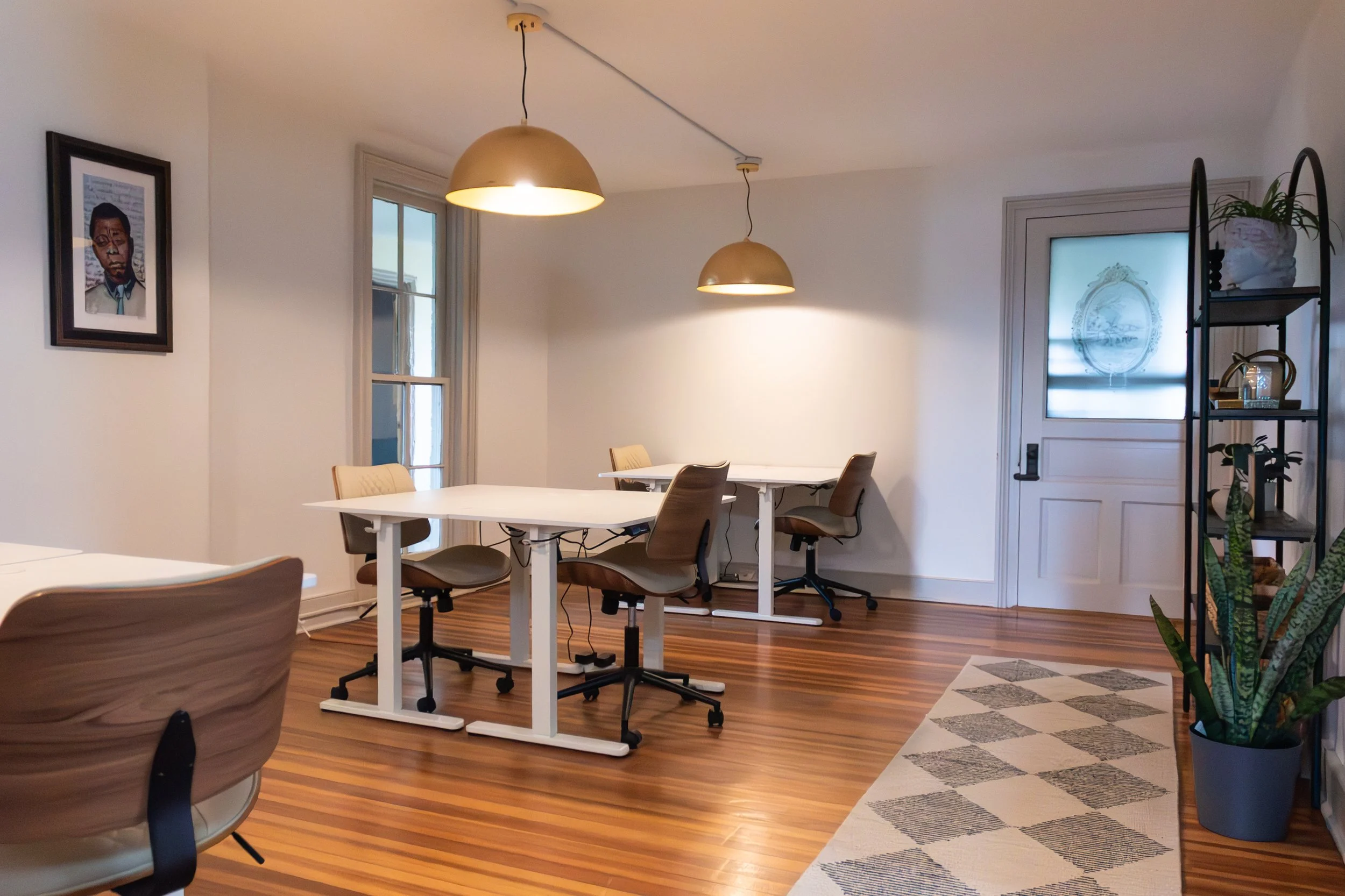 Interior of a modern room with white walls, wooden floor, multiple tables and chairs, a wall art portrait, a shelving unit with plants, and hanging gold-colored pendant lights.