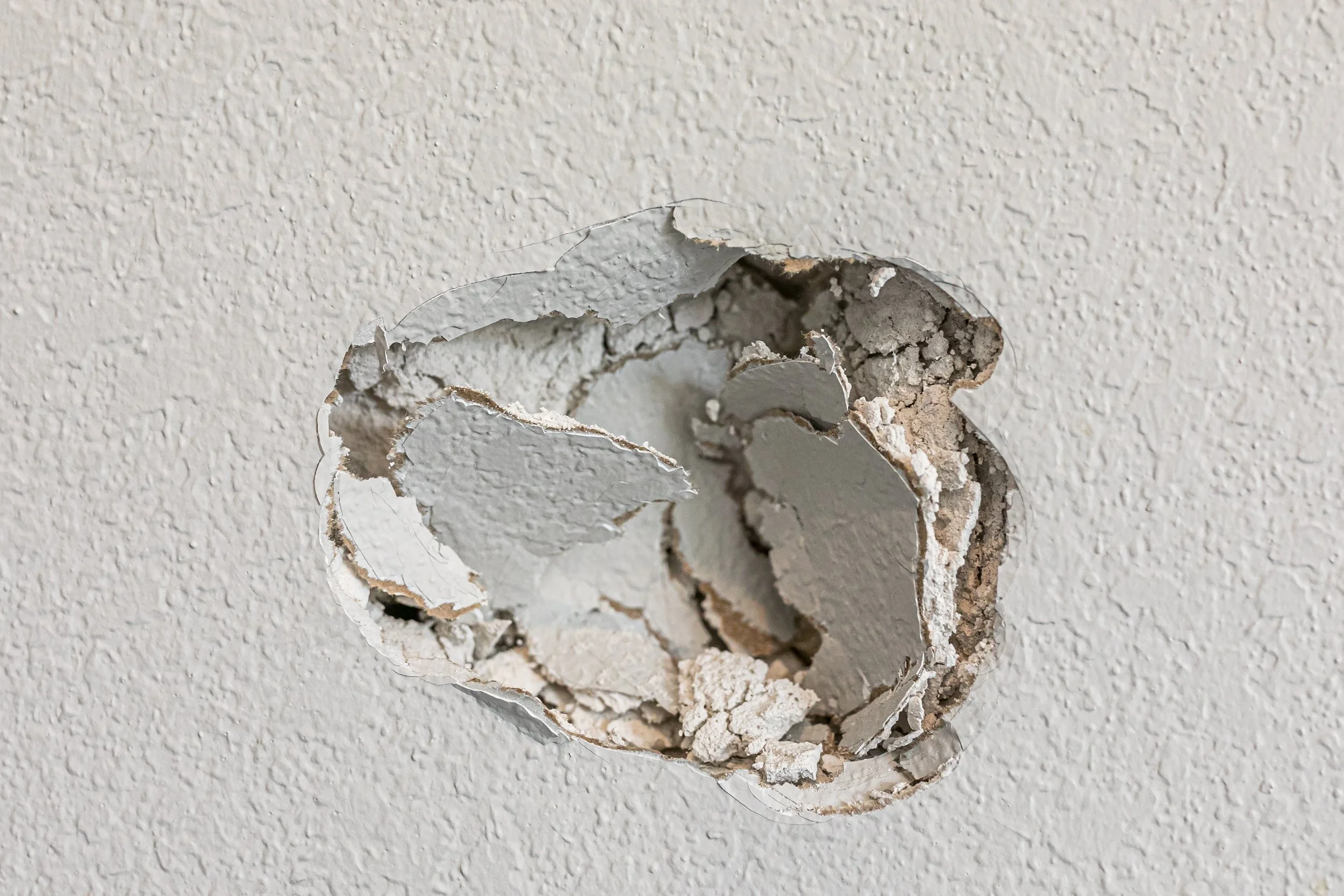 Close-up of a hole in a white textured wall, showing broken drywall and exposed material underneath.