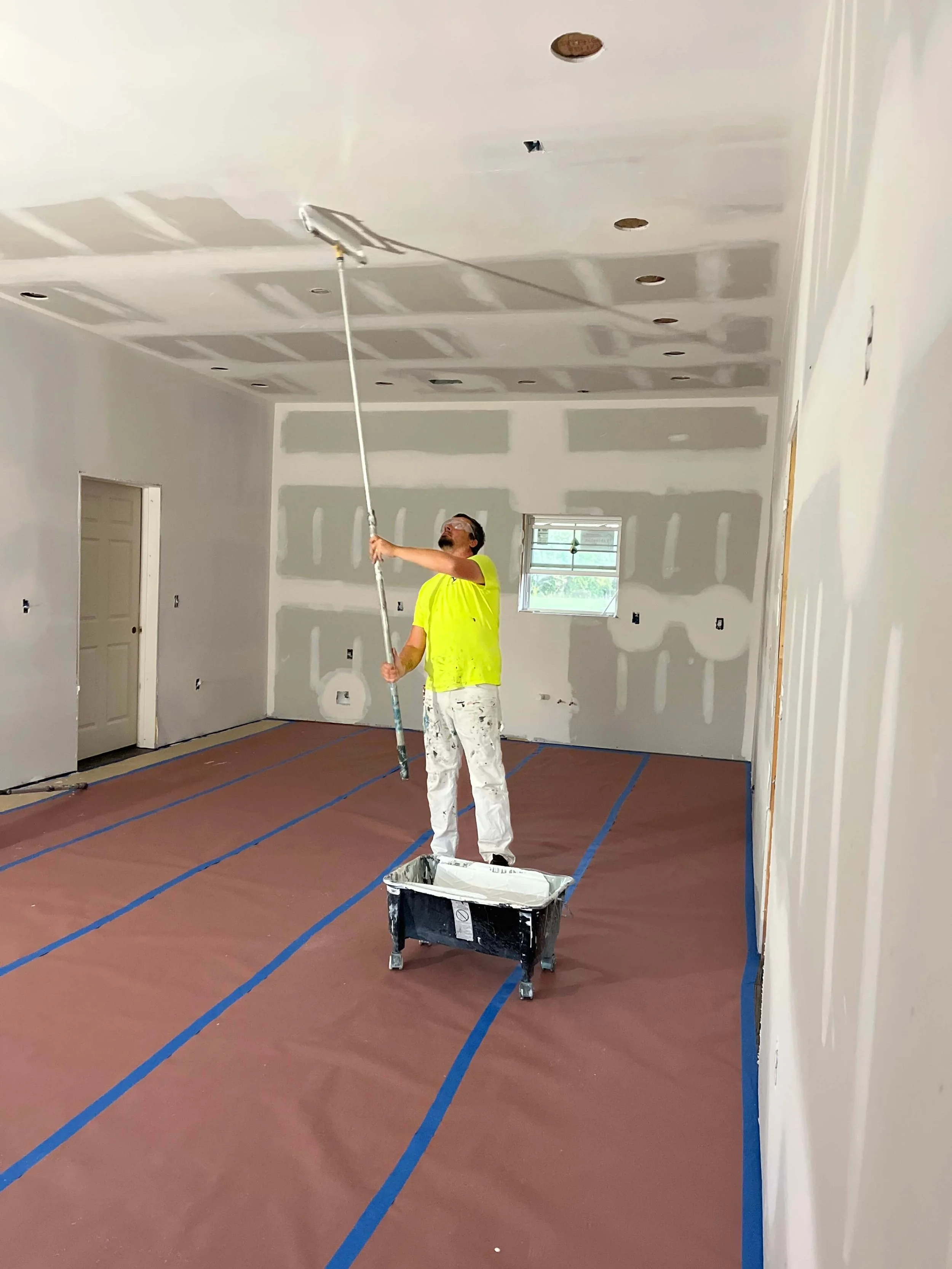 A man in a yellow t-shirt and white paint splattered pants is painting a ceiling with a paint roller. The room has drywall with visible joint compound, and painter's tape is on the floor and wall edges. A trash bin is in front of him, and a window is in the background.