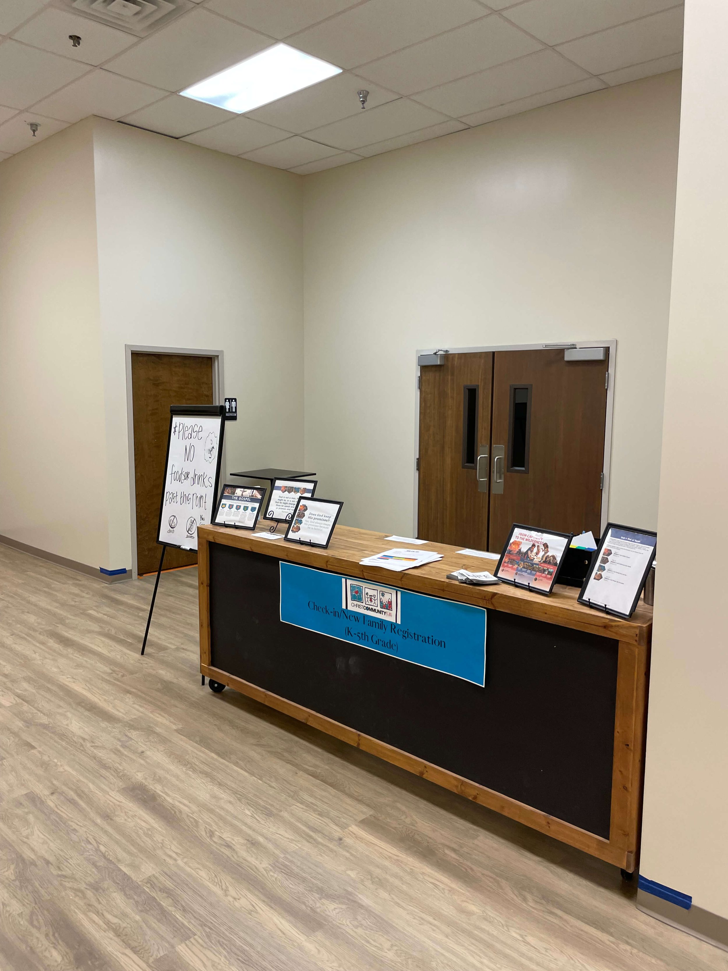 A registration table with signs and papers in a room with beige walls and wood-like flooring. The main sign on the table reads 'Check-in/NEW Family Registration (K-5th Grade).' Behind the table, there are framed informational signs, and to the left, 