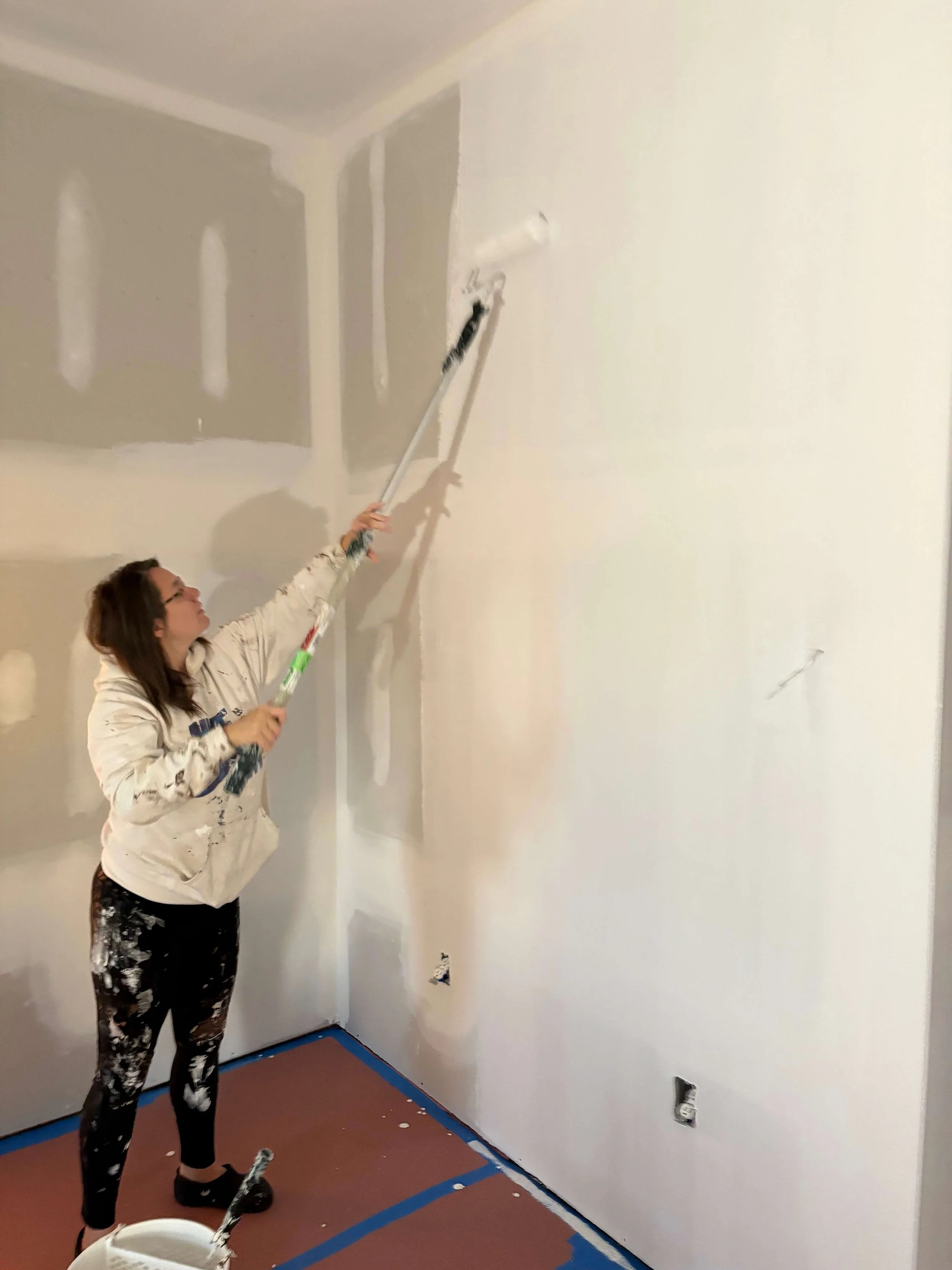 A woman painting a wall with a roller in a room that is being renovated, with drywall and unfinished surfaces visible.