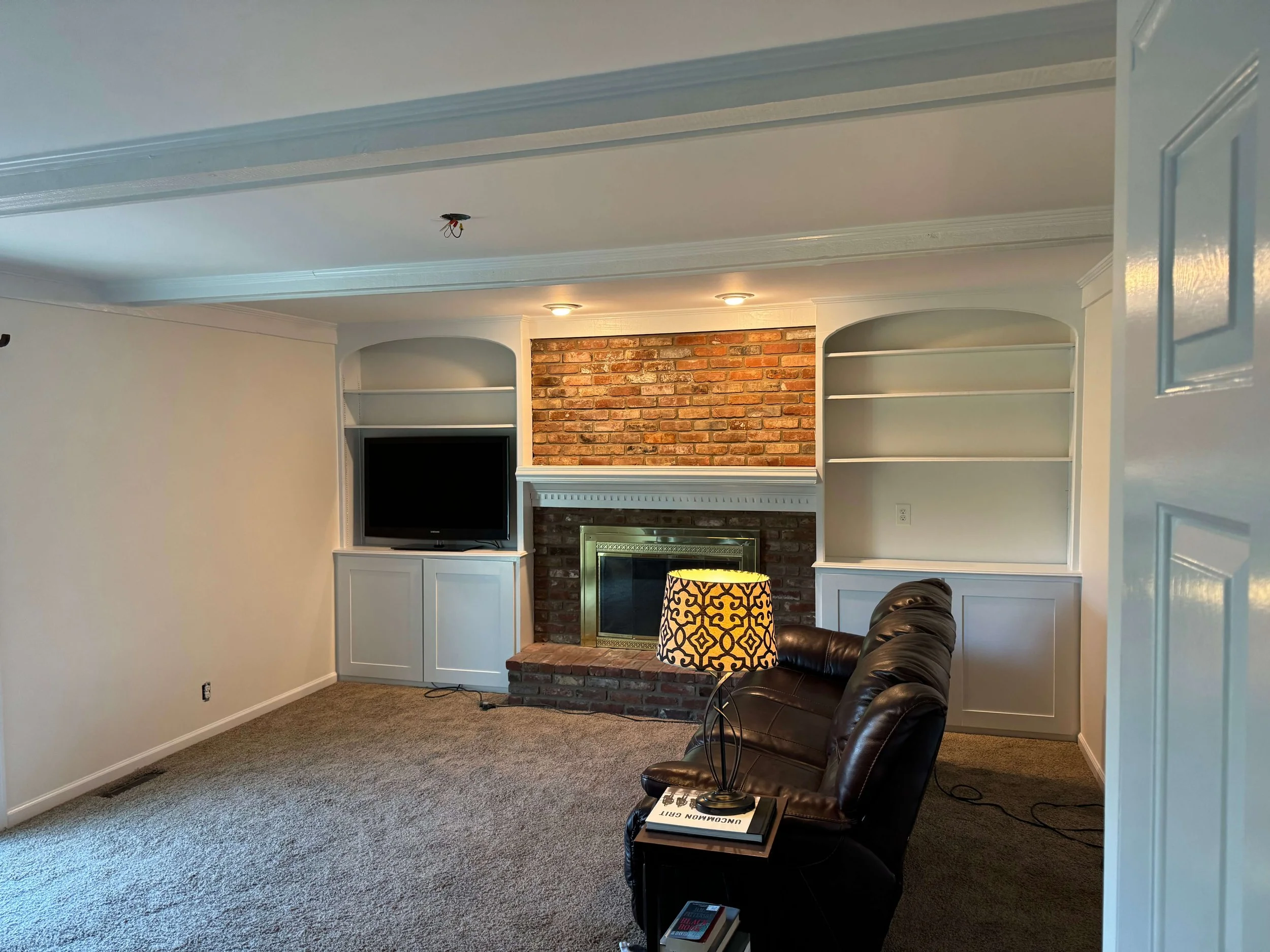 Living room with a brick fireplace, white built-in shelves, a TV, a brown leather sofa, a table lamp with a patterned shade, and books on the floor.